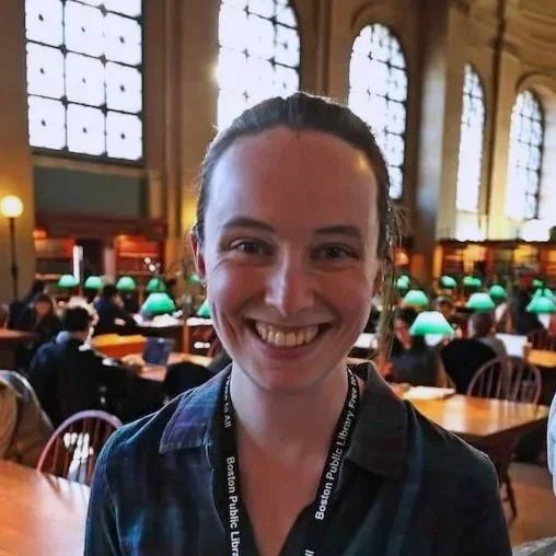 Woman smiling in a large, historic library filled with wooden tables, green banker lamps, and large arched windows.