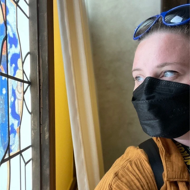 Woman with glasses on her head and a black face mask gazing to the right, beside a stained-glass window and a yellow wall, and wearing a burnt orange shirt.