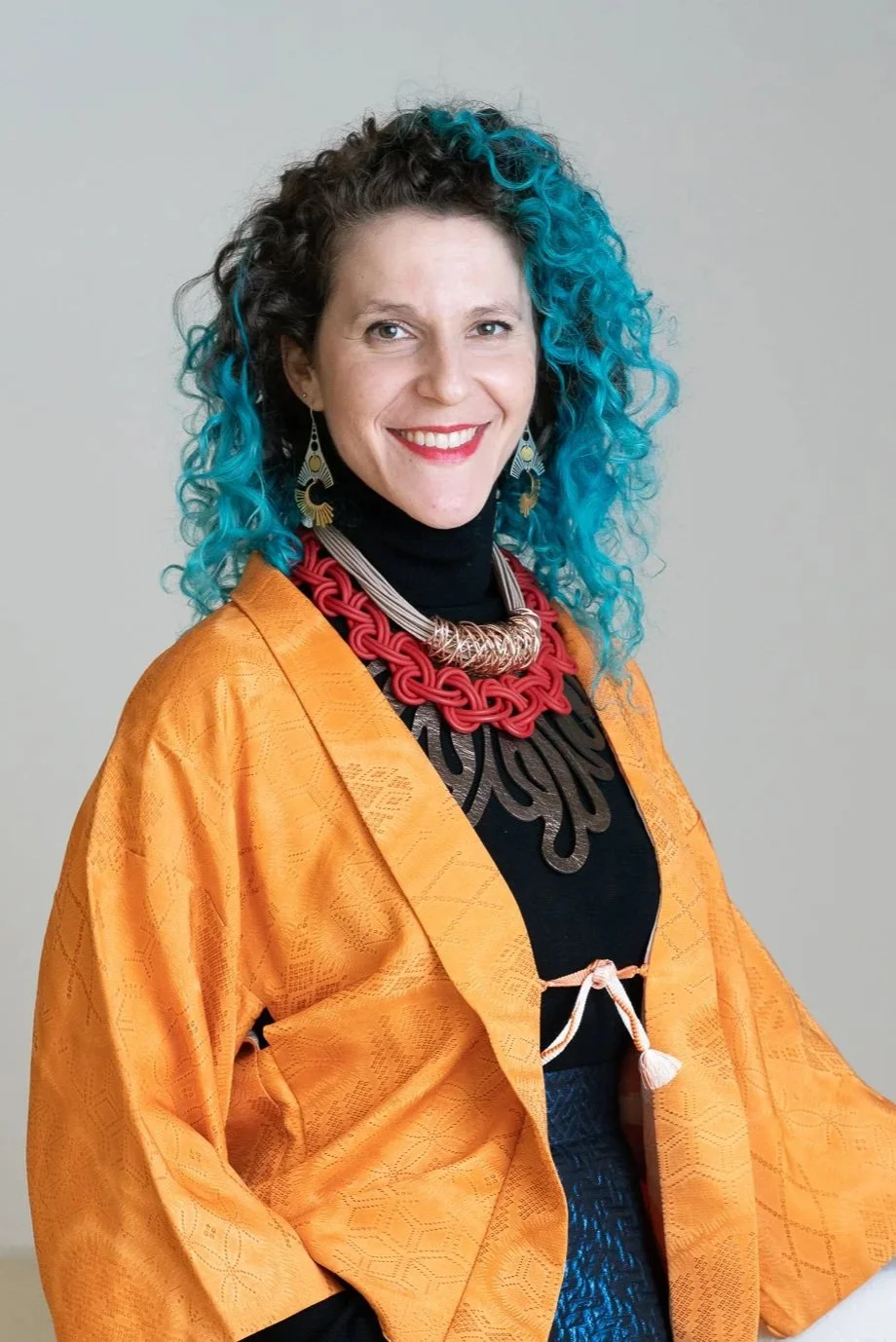 A woman with curly hair that is half black and half bright blue, smiling at the camera. She is wearing a black turtleneck, a vibrant orange jacket, and bold jewelry including large earrings, a red chunky necklace, and other layered necklaces.