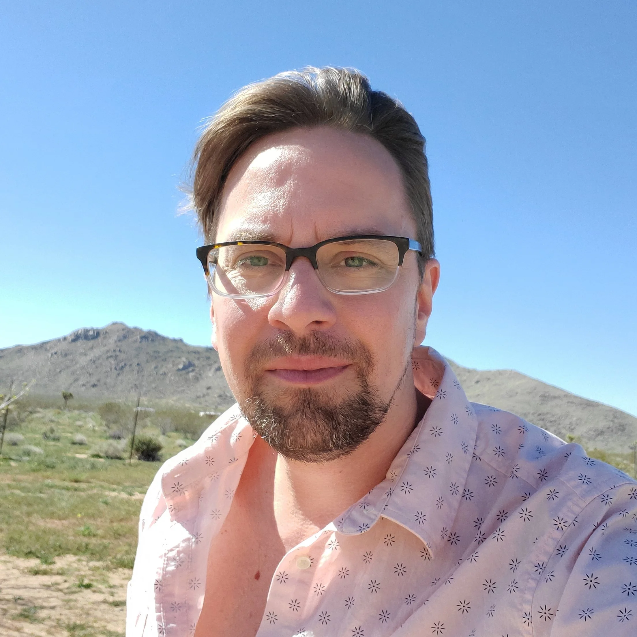Selfie of a man with light brown hair, a short beard, and glasses, wearing a light pink floral shirt. The background features a mountain range under a clear blue sky.