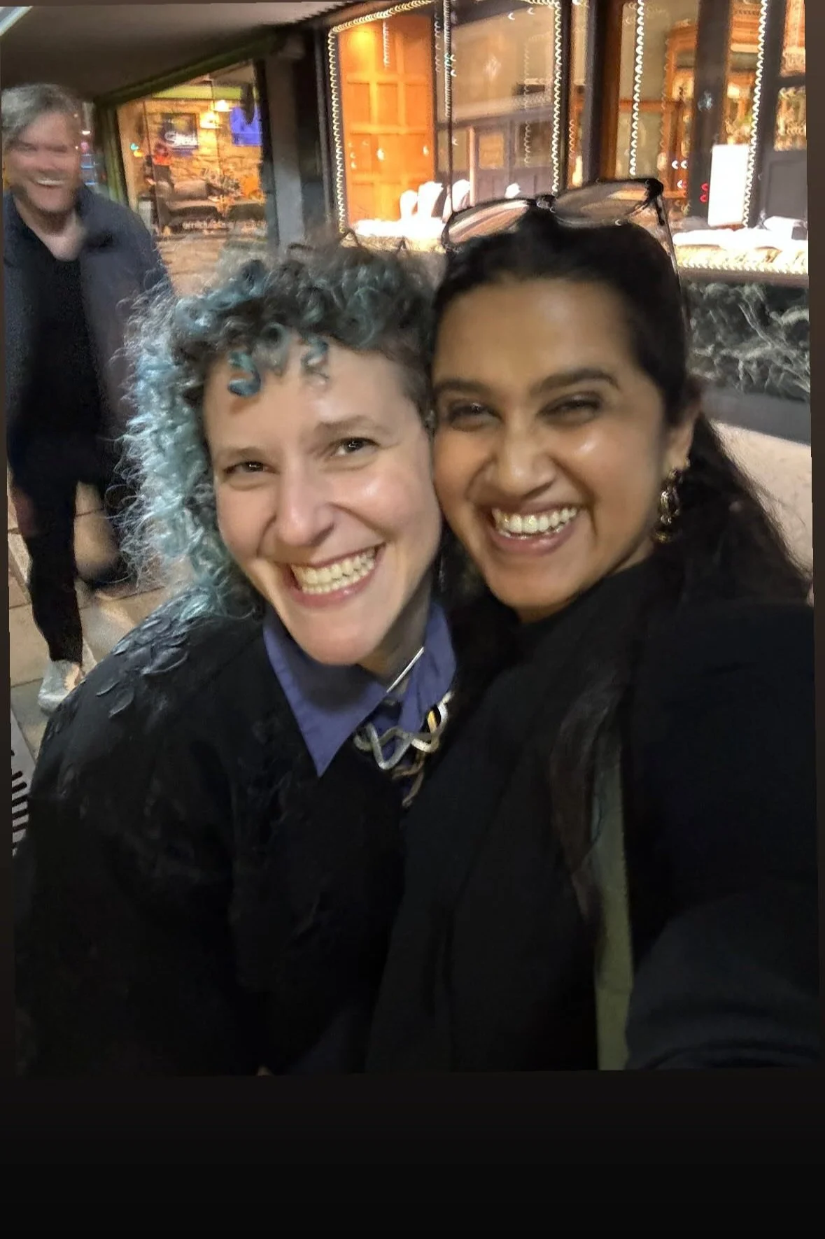A selfie of a toothily grinning white-skinned woman and brown-skinned woman in the foreground while a blurry, also-grinning white-skinned man photobombs them in the background.