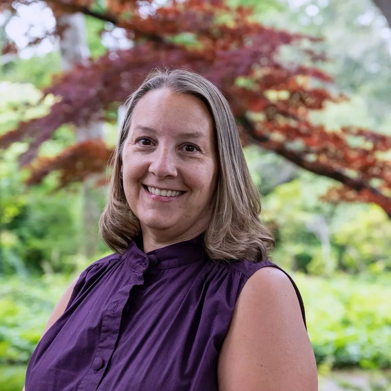 A smiling woman with shoulder-length hair posing outdoors with a red-leafed tree and green foliage in the background.