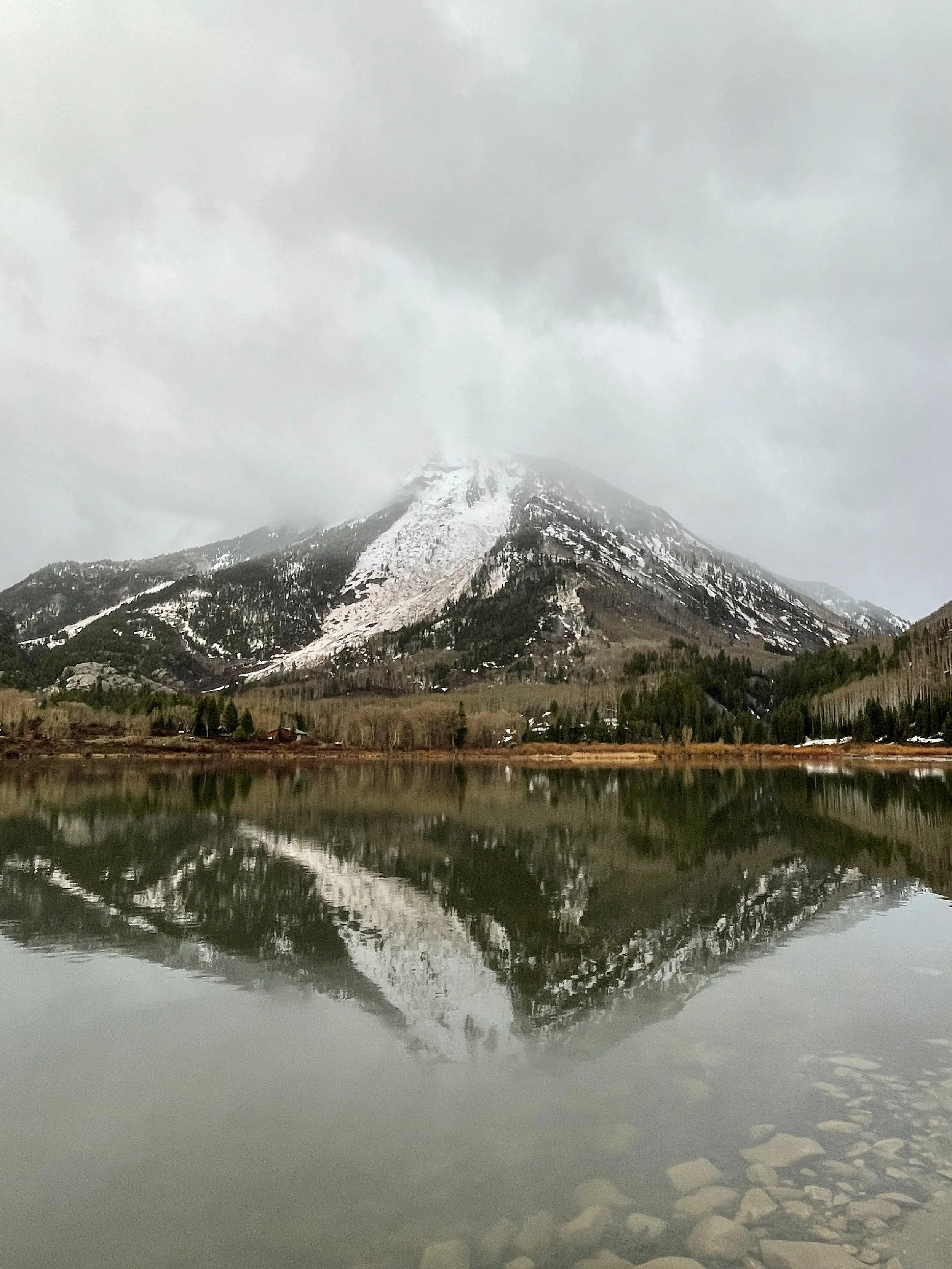 A photo of a snow-sprinkled mountain covered in trees and reflected in the pond below it. The sky is misty and full of gray foggy clouds.