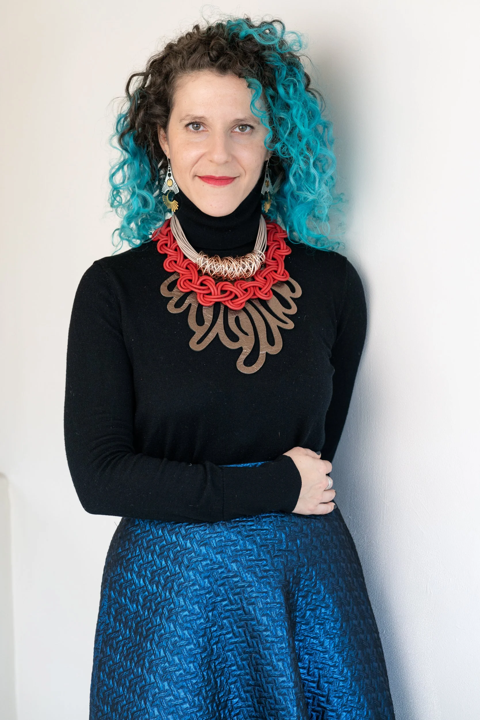 A person with curly, blue-tinted hair wearing a black turtleneck, layered red and tan sculptural necklaces, and a shimmering blue textured skirt.
