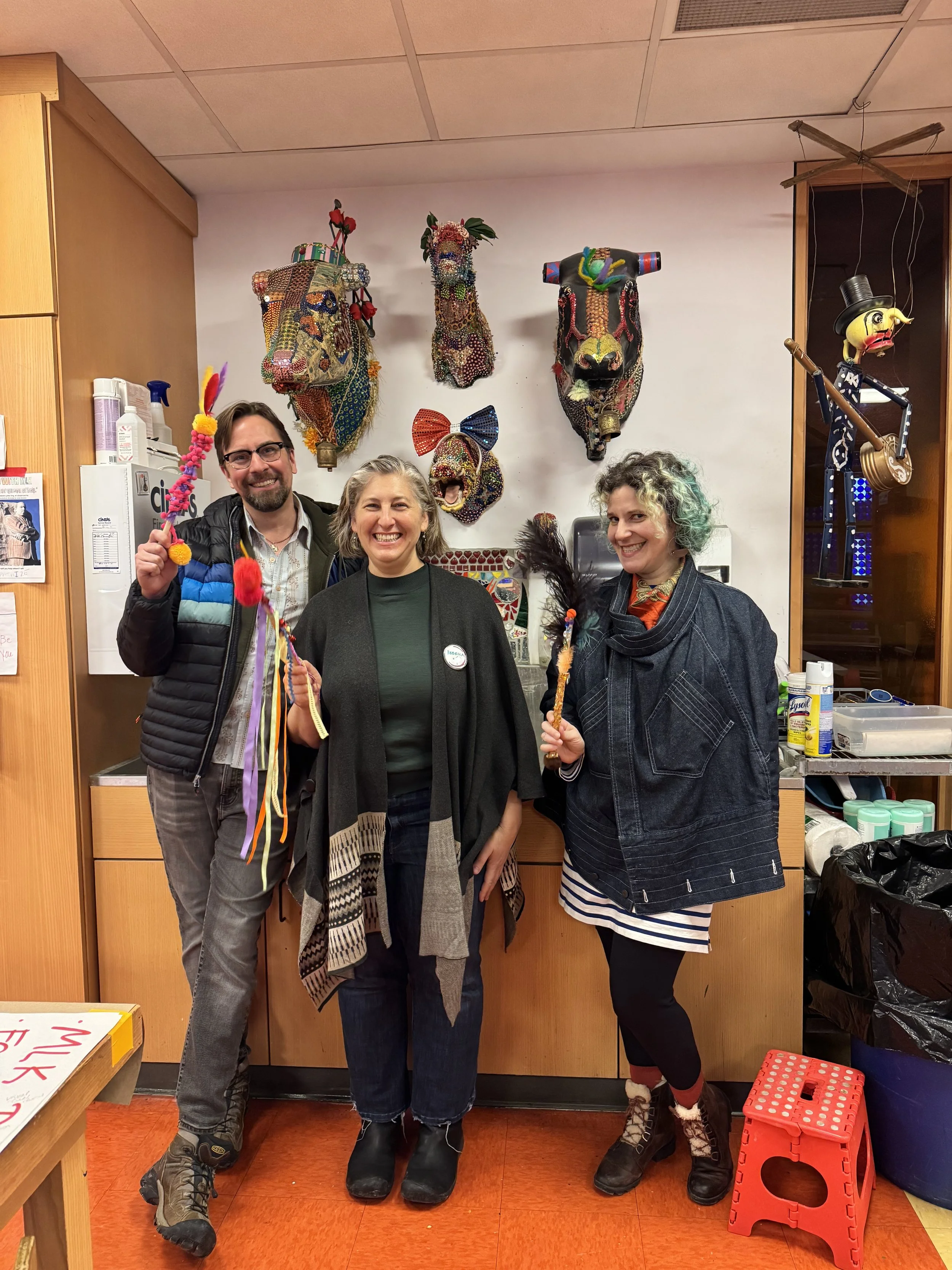 Three people smiling and holding decorated wands, posed in an art studio, with various art pieces adorning the walls, including ornate masks and a puppet.