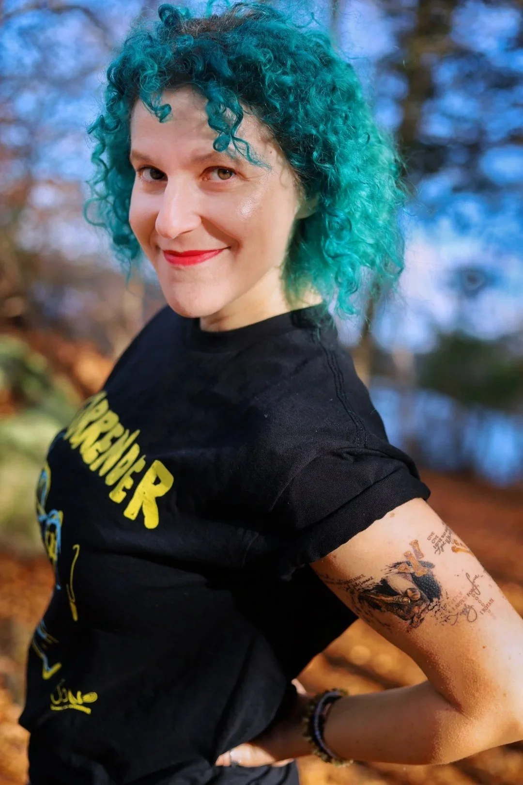 A woman with curly teal-colored hair smiling outdoors during fall, wearing a black T-shirt with yellow text and displaying tattoos on her left arm.