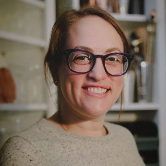 A woman with red hair, wearing glasses and a beige sweater, smiling in a room with shelves in the background.
