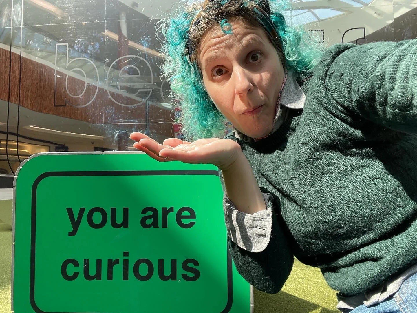 A person with teal-colored hair makes a playful shrugging gesture towards the camera. They are positioned next to a bright green sign with black text that reads "you are curious."