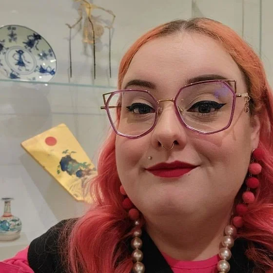 A woman with pink hair, glasses, and red lipstick smiles while posing in front of a display case filled with artifacts.