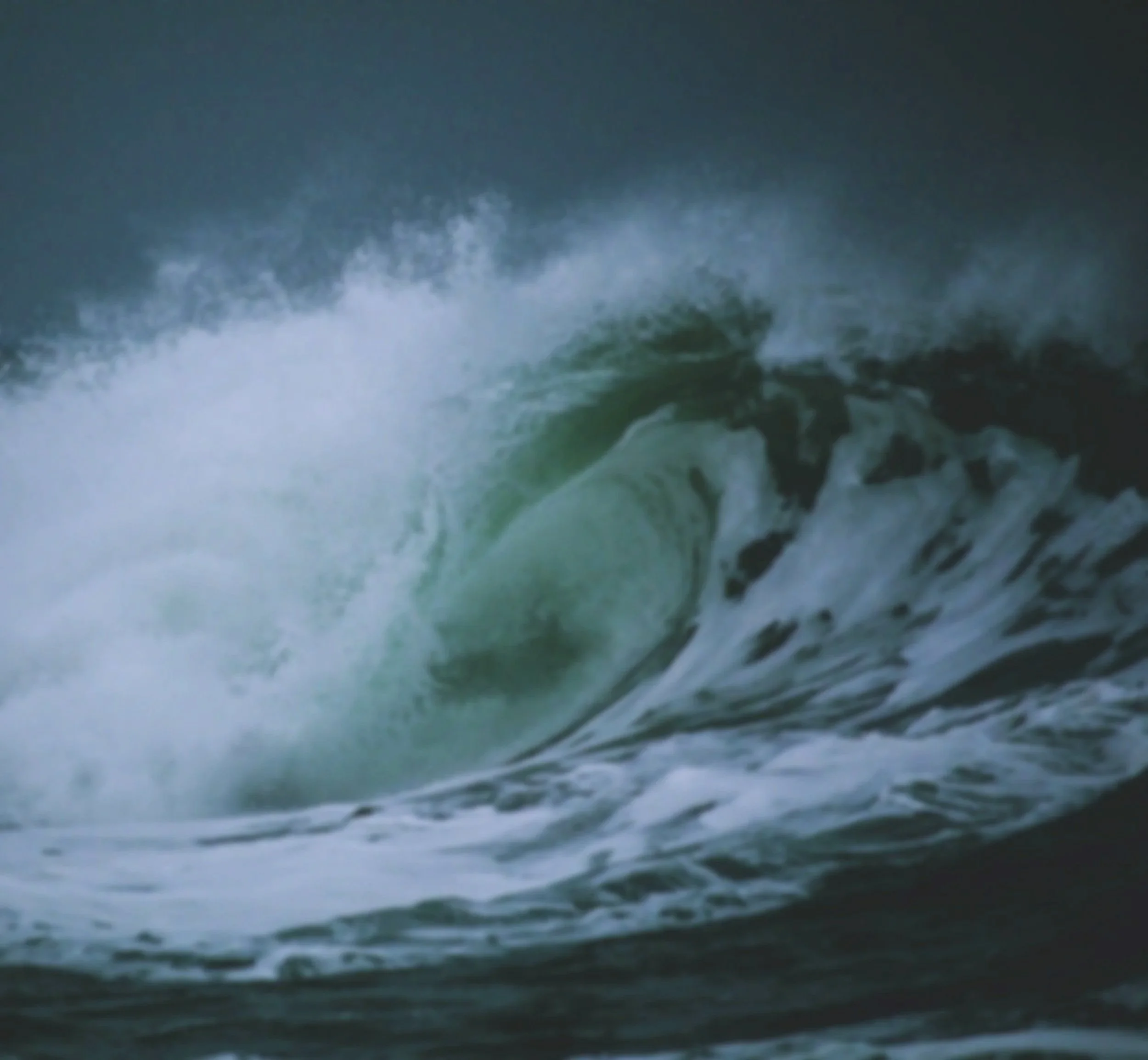 A large ocean wave curling and crashing with white foam at the crest, dark stormy sky in the background.