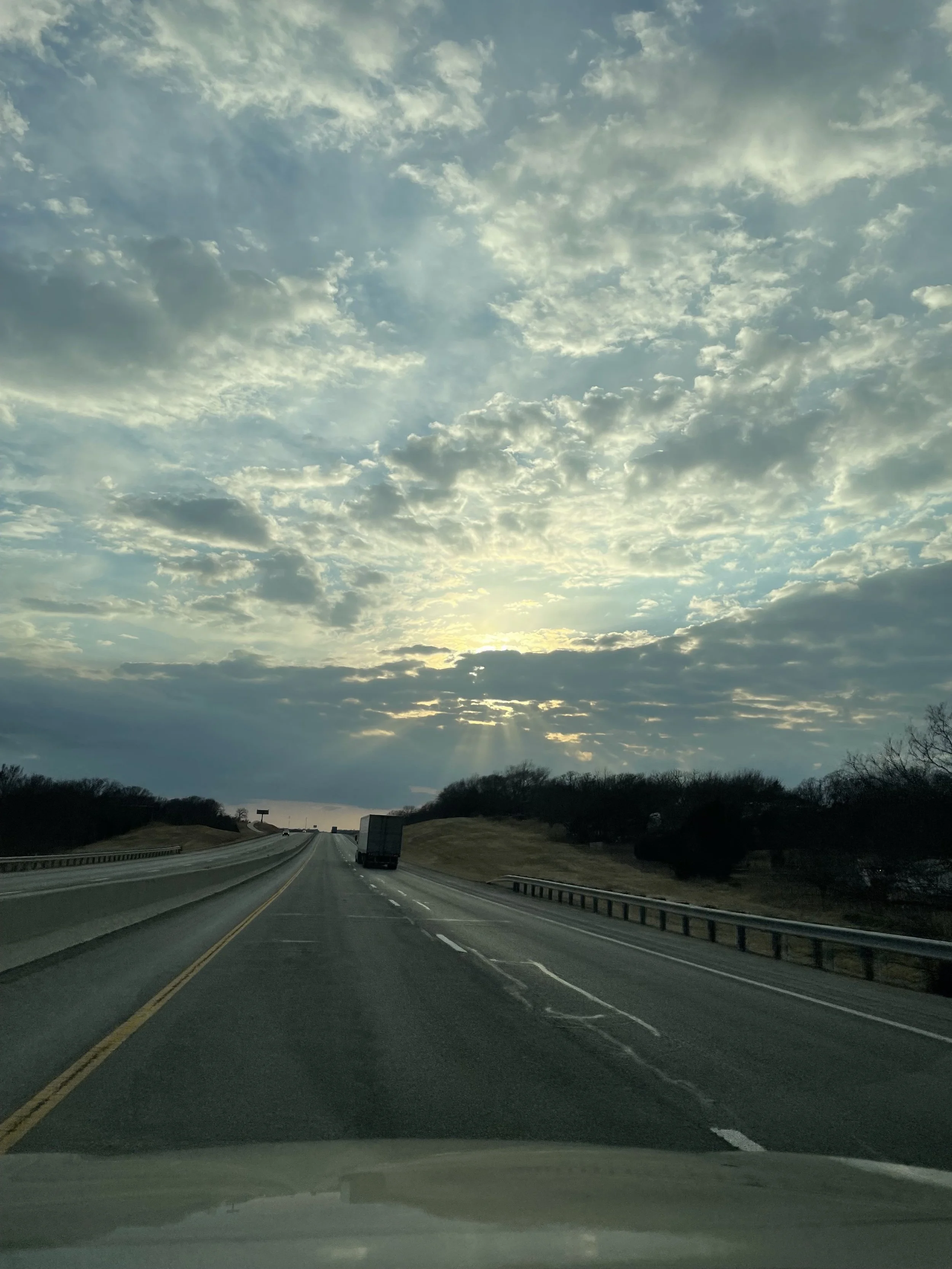 A photo of a wide expanse of blue sky covered with patchwork of white clouds that are highlighted bright around their edges. The sun is setting behind some clouds low at the horizon and rays of streaking light can be seen descending to the highway.