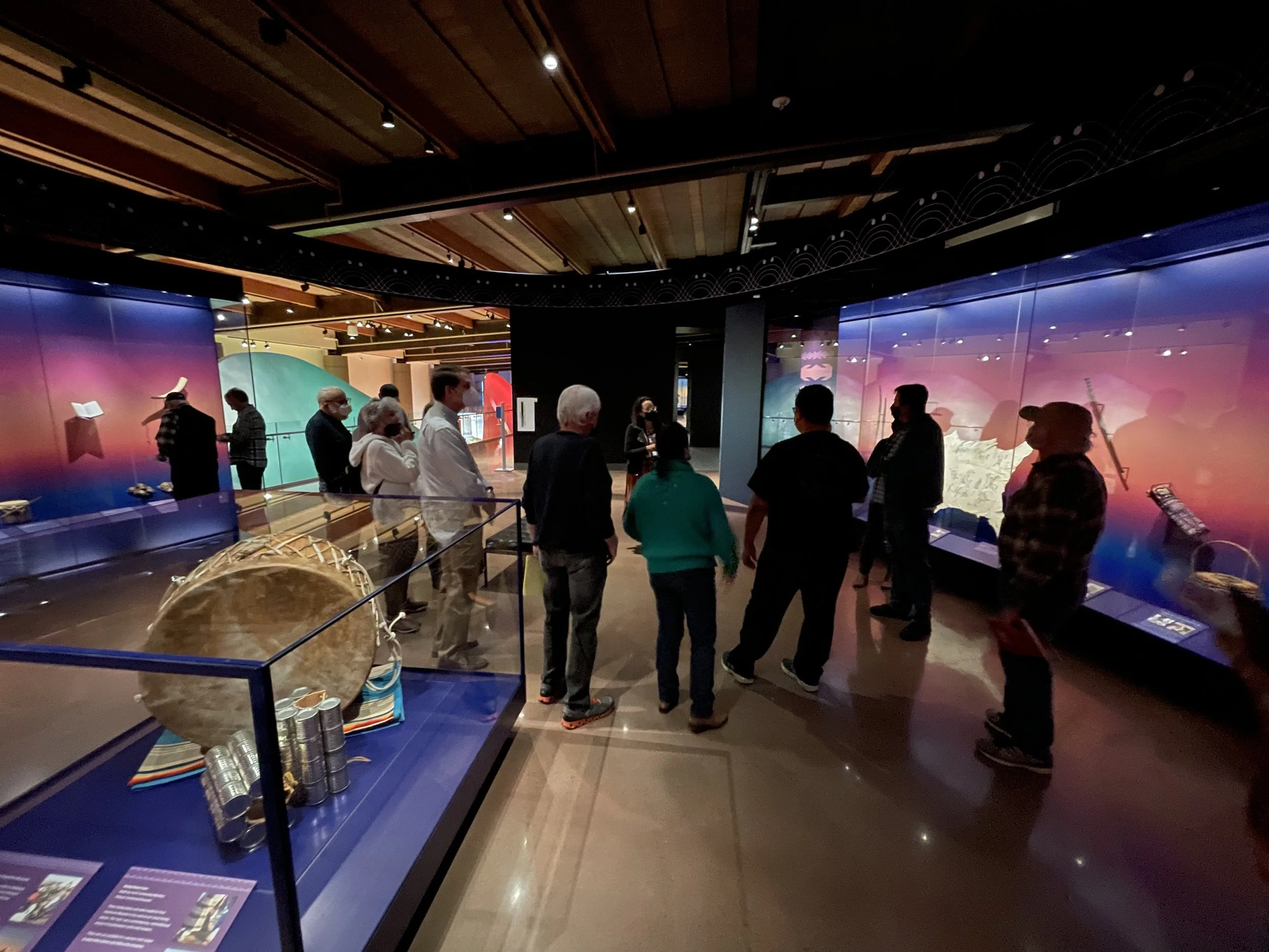 A photo of Adrienne leading a tour for her group of docents who are arranged around her in an exhibition space that's got sunset-colored wall colors on each side and a case with a large drum and leg shakers in the foreground.