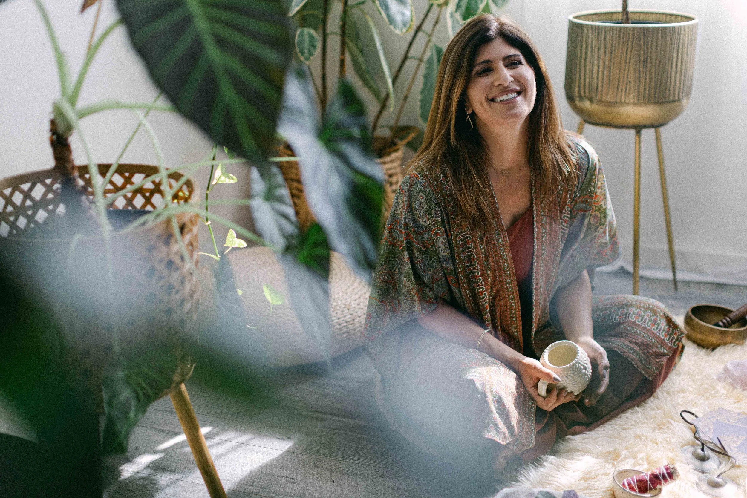 A woman sitting cross-legged on the floor among indoor plants, smiling and holding a decor object, with a floor lamp nearby and various decorative items around her.