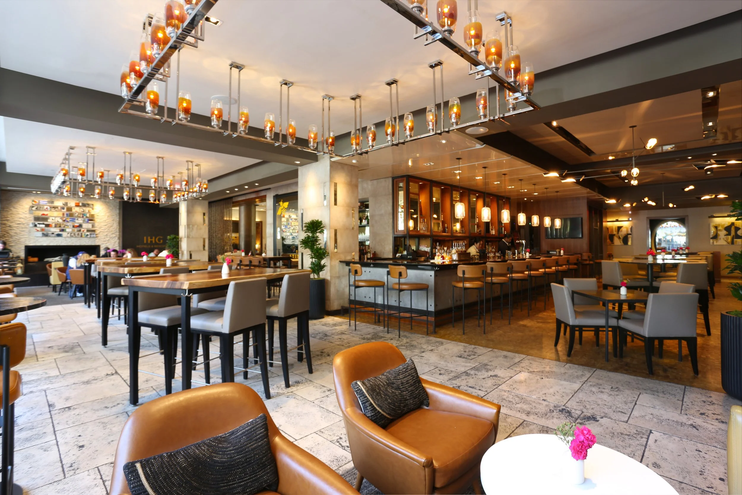 Interior of a modern restaurant or bar with various seating options, pendant lighting, and a bar area with barstools and liquor shelves.