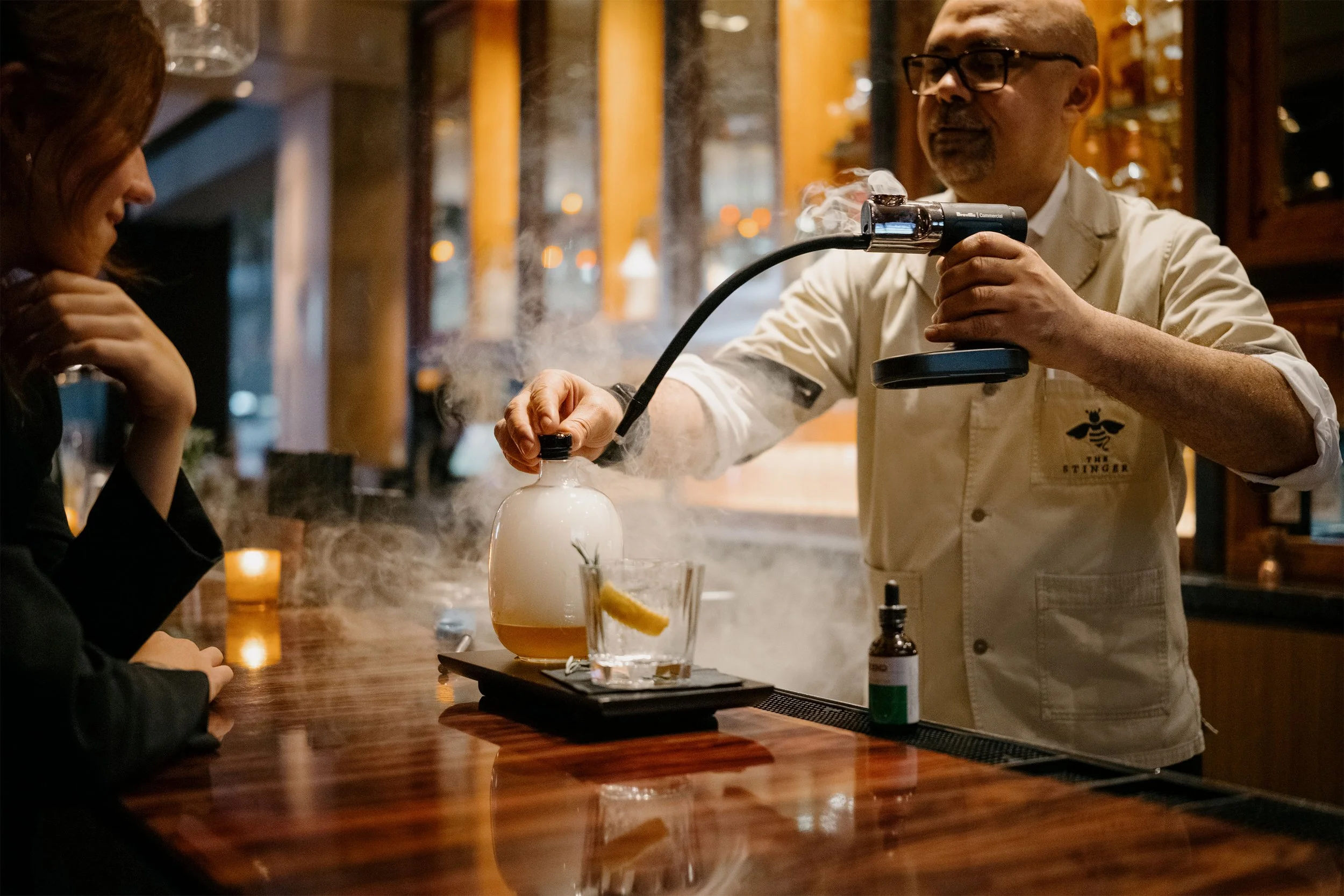 Bartender in beige shirt performing a flameless distillation or smoke presentation with a science apparatus at a bar, while a woman watches.
