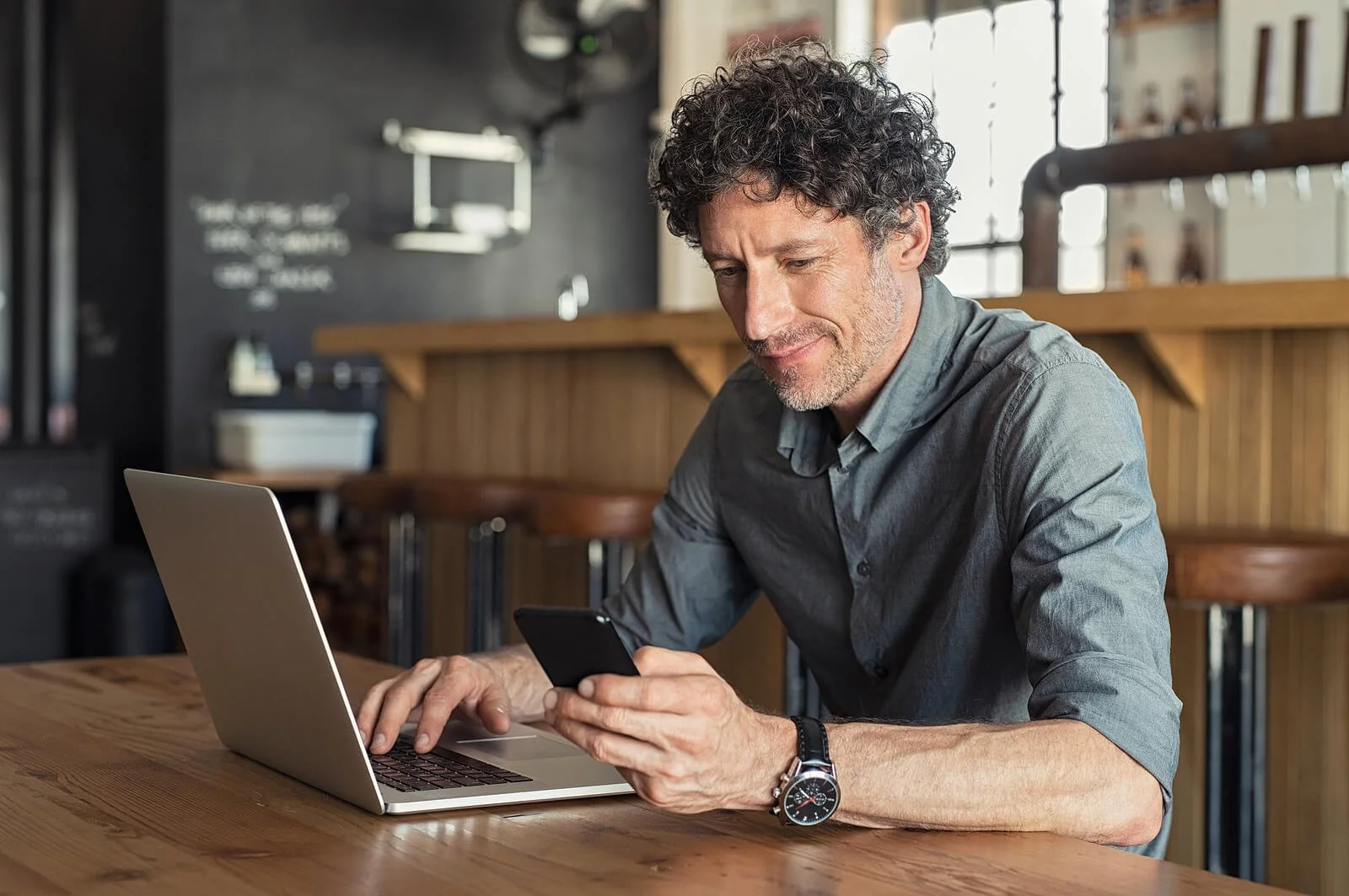 Middle-aged man with curly hair smiling at his phone while working on a laptop at a wooden table in a cafe. Knowing what to do when your Google Ads for Therapists account is compromised can save your budget and keep your lead flow intact.