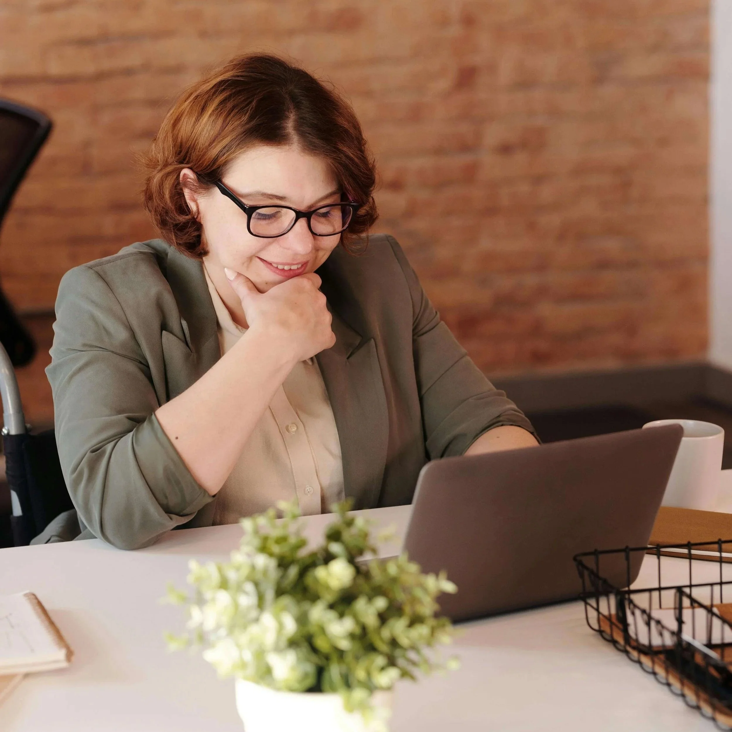 Woman wearing glasses smiling while working on laptop at desk with brick wall and plants. Your website is the digital front door that determines if Google Ads for Therapists clicks become booked clients.