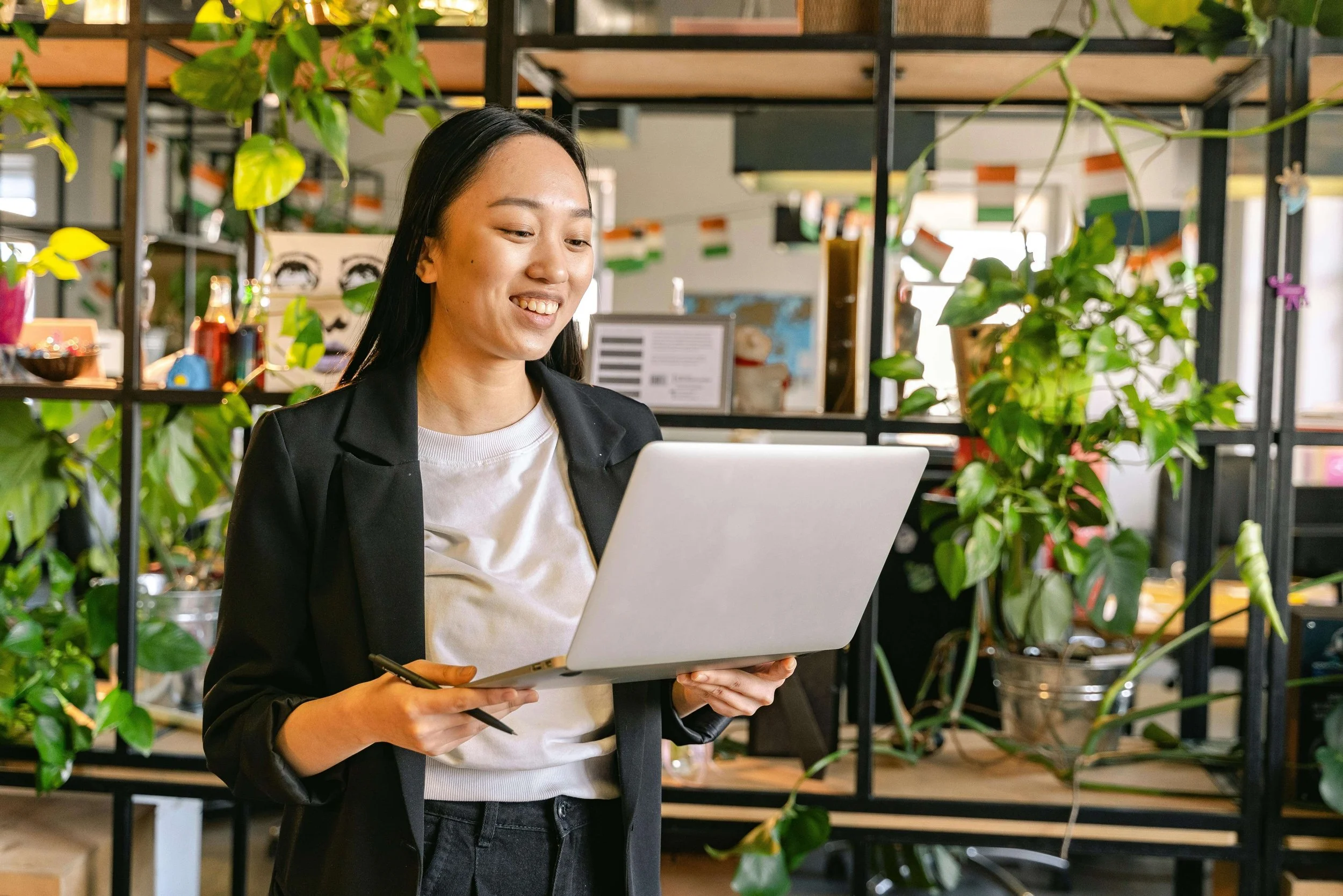 Woman in black blazer smiling while holding laptop in modern office with plants and shelving. Understanding seasonal trends helps you manage Google Ads for Therapists budgets during summer slowdowns and fall surges.