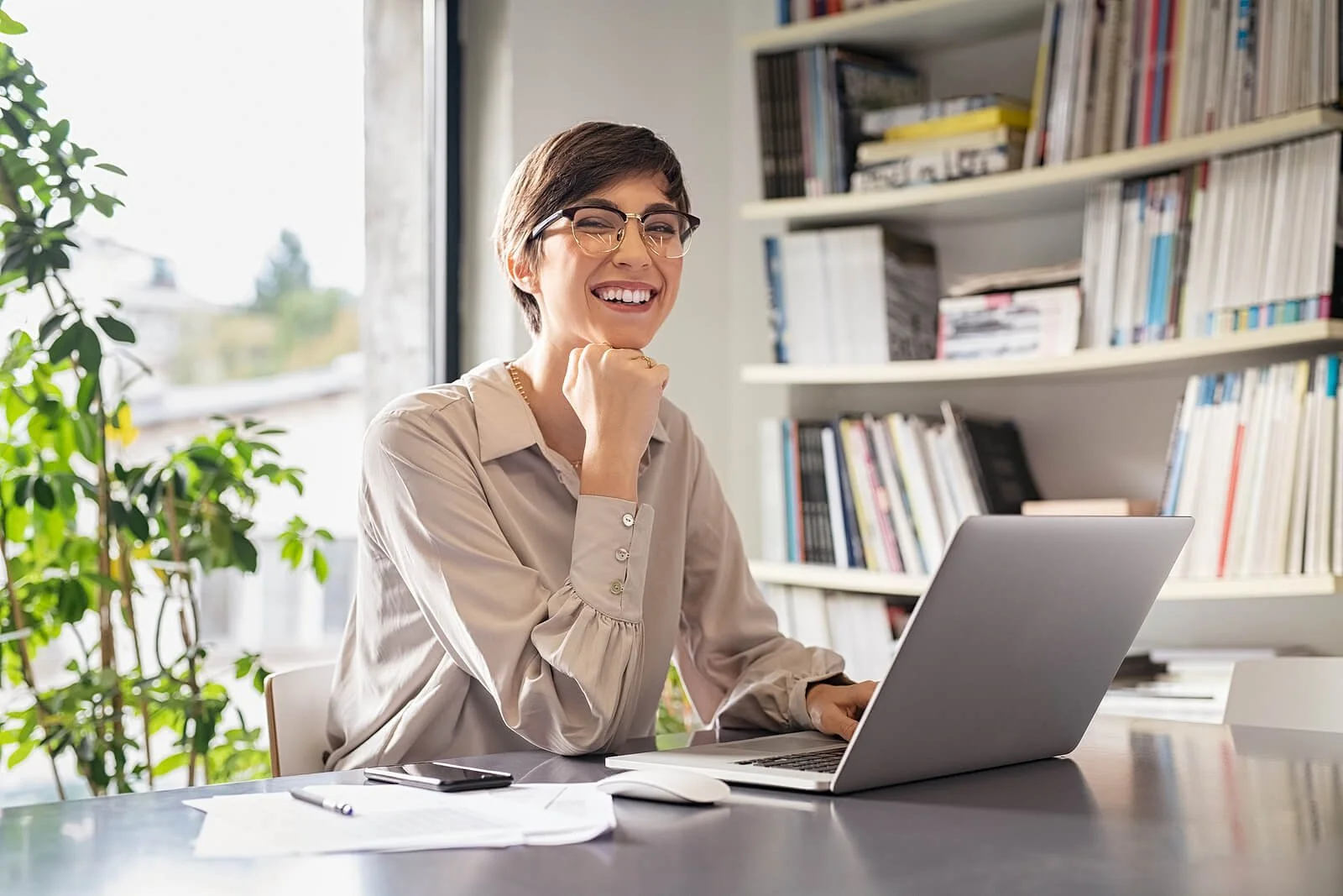 Woman wearing glasses smiling at laptop with bookshelf and plant in bright office. Understanding the 90-day timeline helps set realistic expectations for Google Ads for Therapists campaign success.
