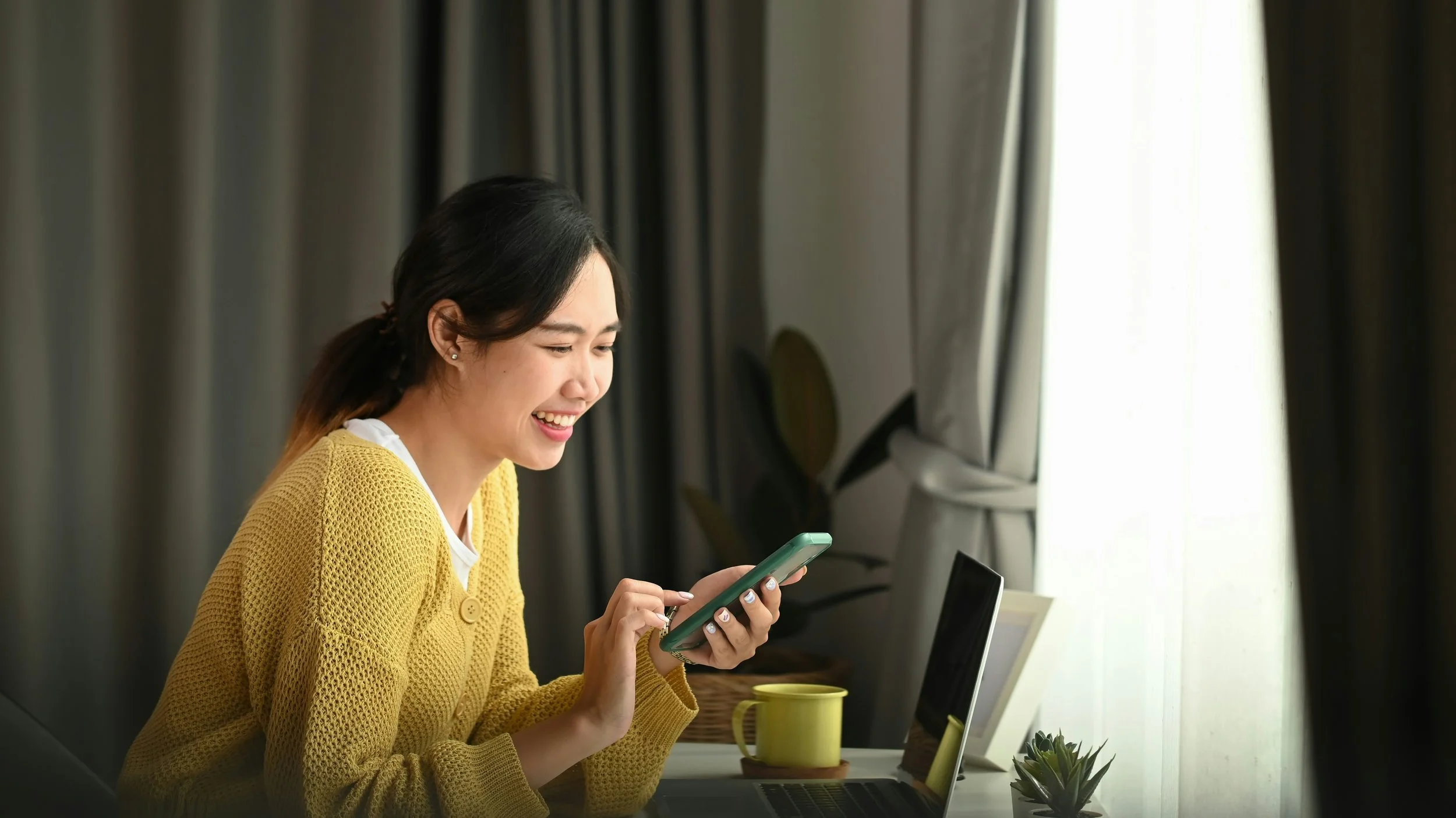 Woman smiling while checking a notification on her phone next to an open laptop