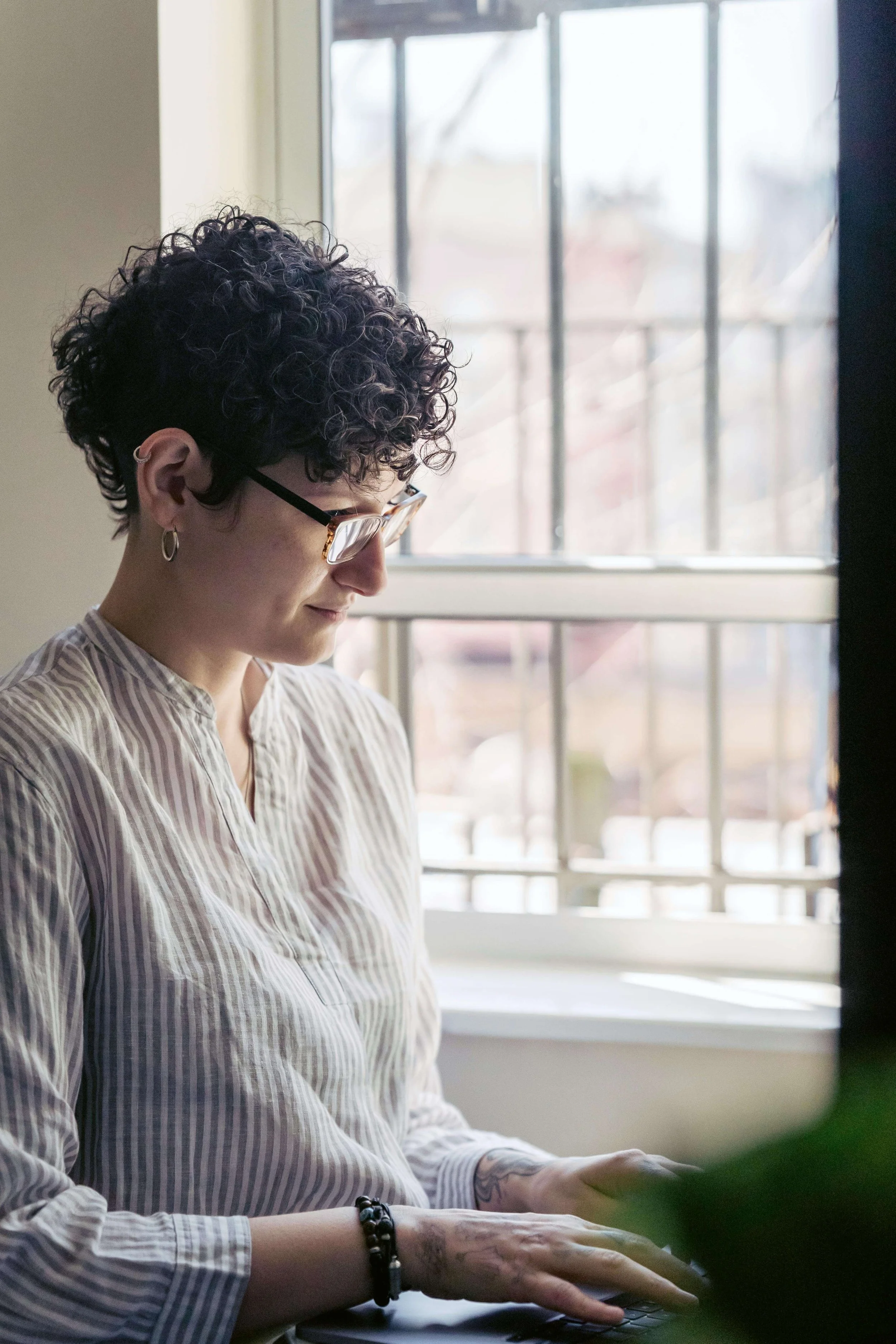 Woman with curly hair and glasses looking thoughtfully out large window with city view. Keep your Google Ads for Therapists running year-round at reduced budgets to avoid costly learning phase resets during slow seasons.