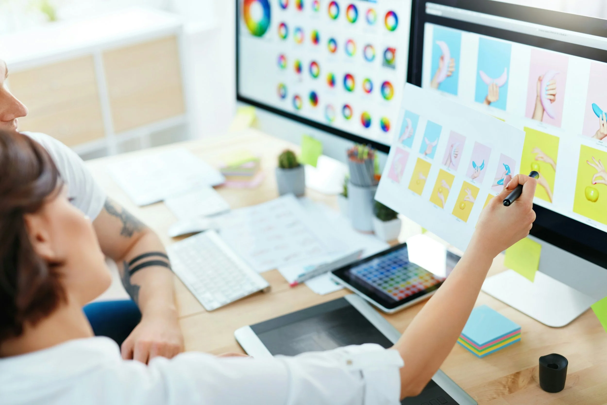 Close-up of two people at a design studio desk comparing a printed sheet of hand-gesture illustrations against a color wheel palette displayed on a monitor, with a tablet and colored sticky notes nearby.