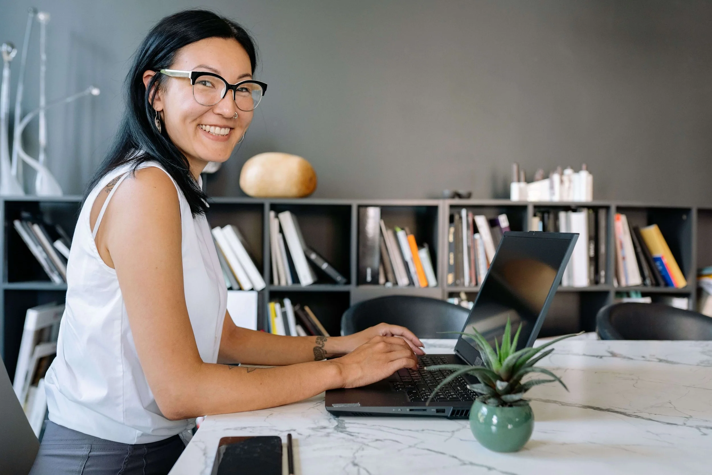 Young woman with dark hair and glasses smiling at the camera while typing on a laptop at a marble desk. Choosing the right Google Ads for Therapists agency starts with knowing the right questions to ask before you sign anything.