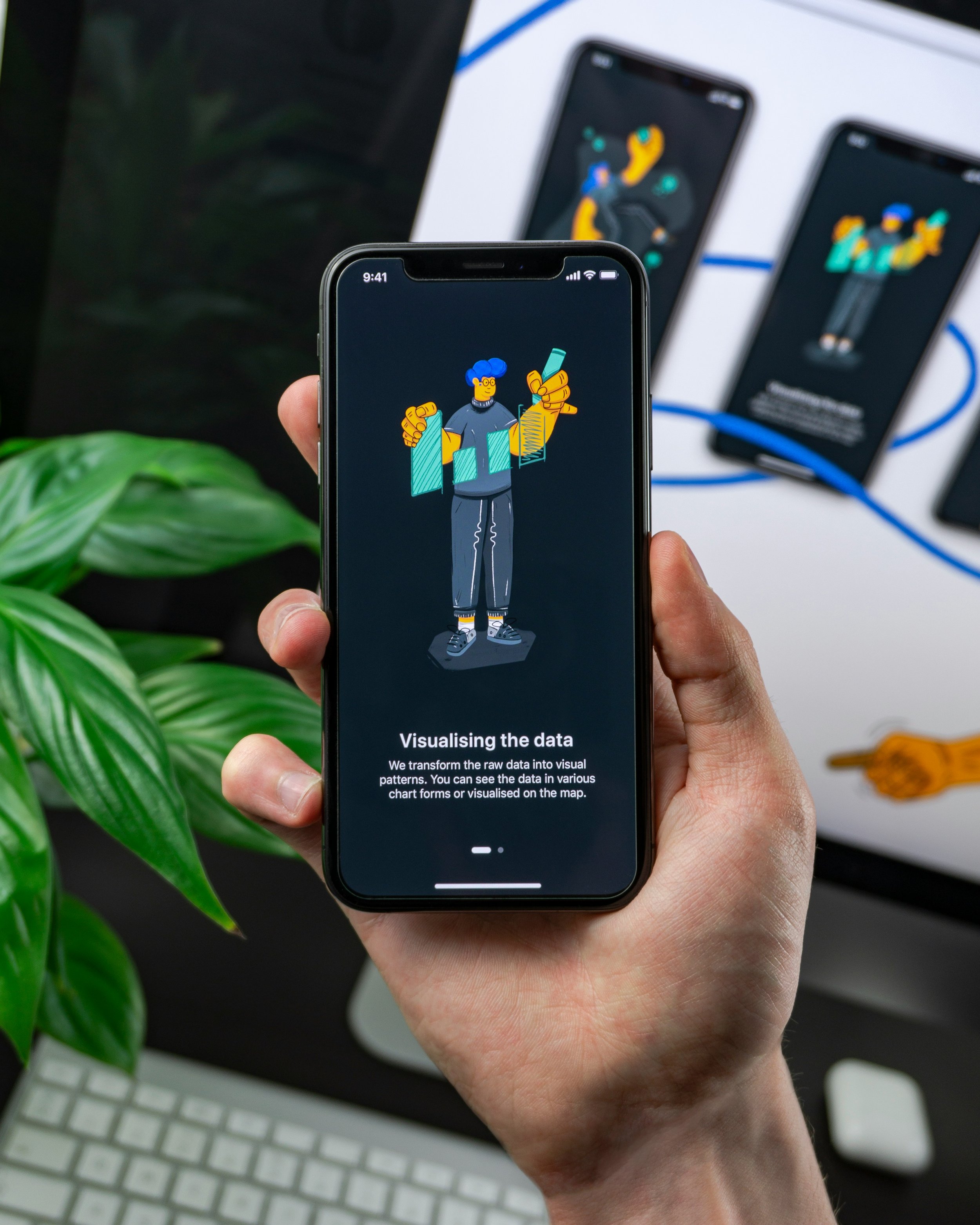 A hand holding a smartphone displaying an app onboarding screen titled 'Visualising the data,' featuring an illustrated character holding chart bars, with a plant, keyboard, and additional phones showing related app screens visible in the background.