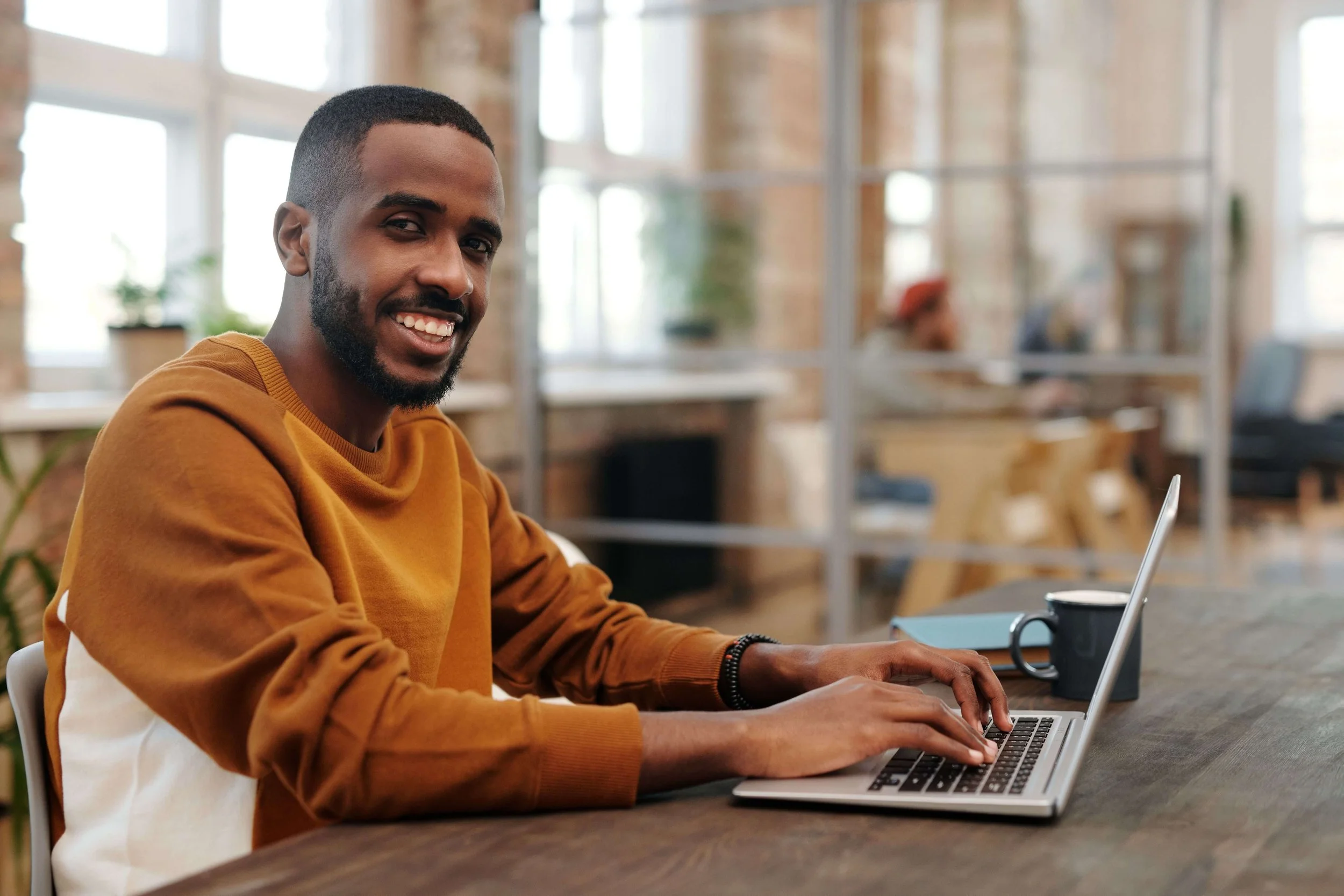 Smiling young man in an orange sweater typing on a laptop at a wooden desk in a bright open workspace. A well-maintained negative keyword list keeps your Google Ads for Therapists budget focused on clients who are actually ready to book.