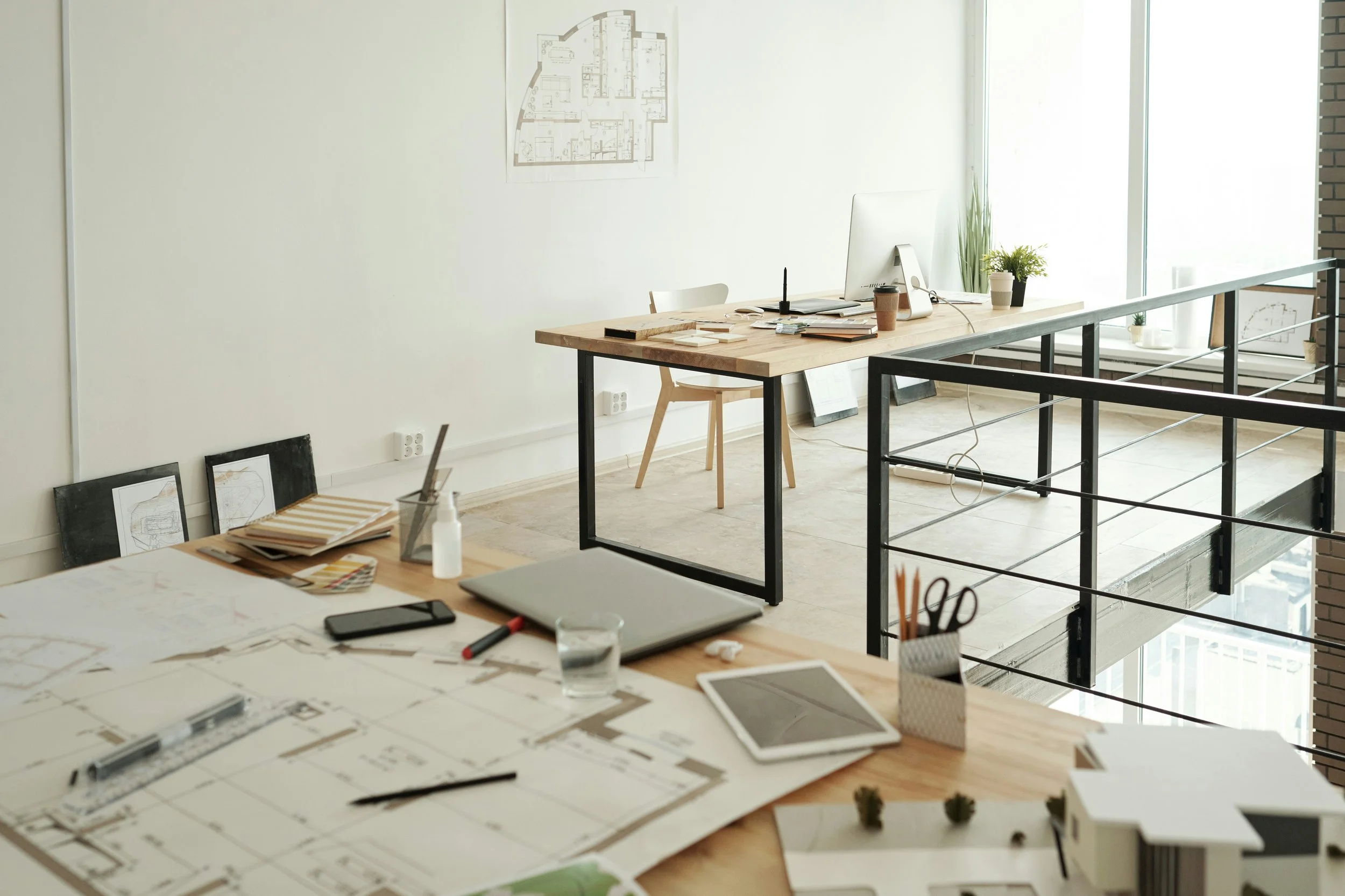 An office with a large window, a desk with a computer, plants, and art supplies, and architectural plans on a table.