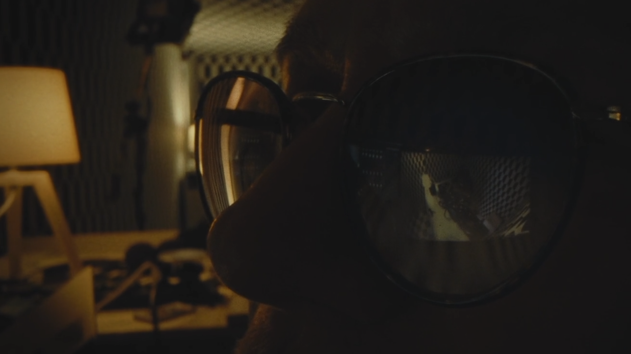 L'image montre une paire de lunettes de protection posées sur une surface sombre, avec un fond intérieur qui inclut une lampe de table et un décor de pièce.