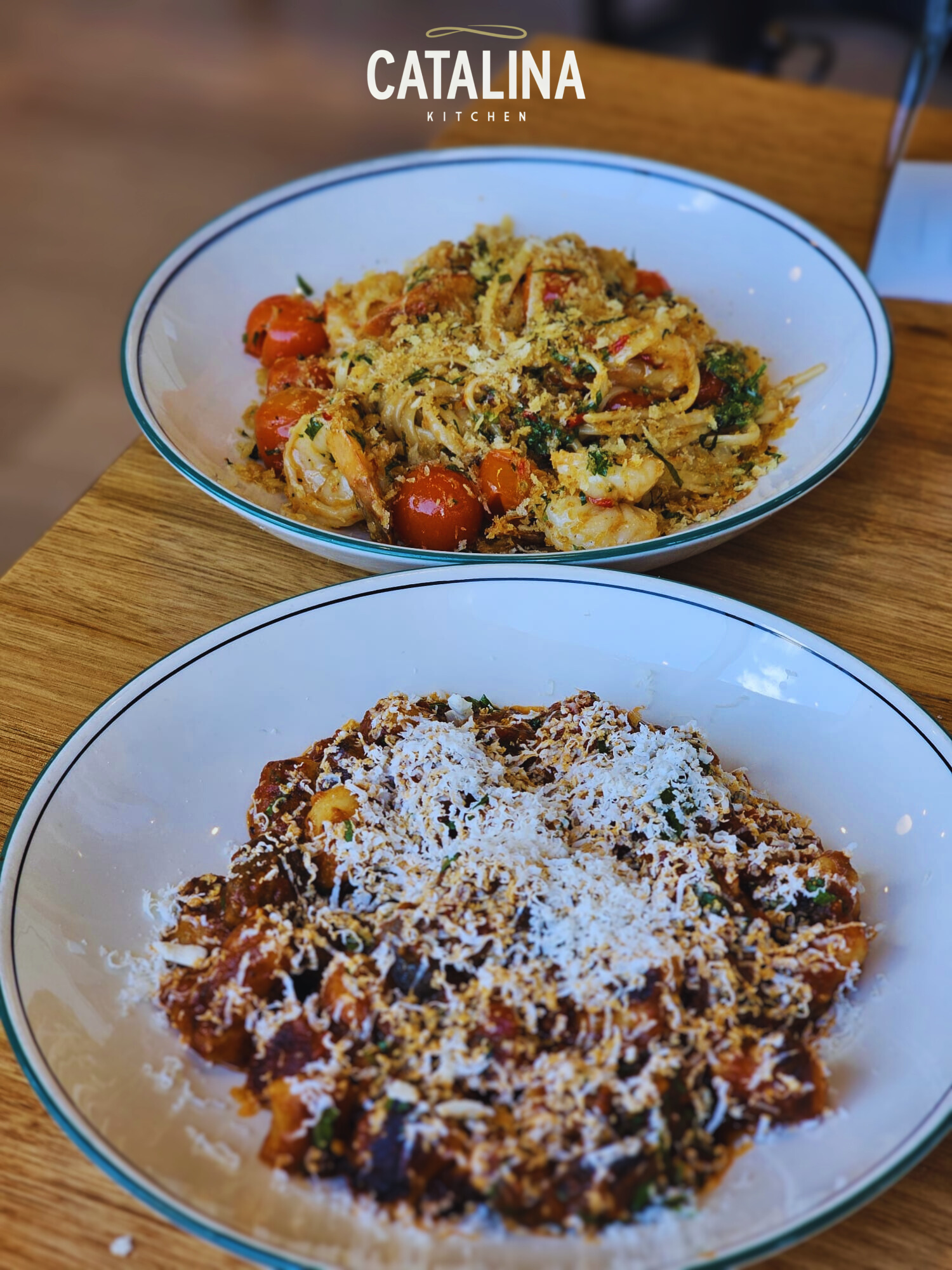 Two pasta dishes in white bowls on a wooden table, one with cherry tomatoes and breadcrumb topping, the other with a tomato-based sauce covered with grated cheese, at Catalina Kitchen.