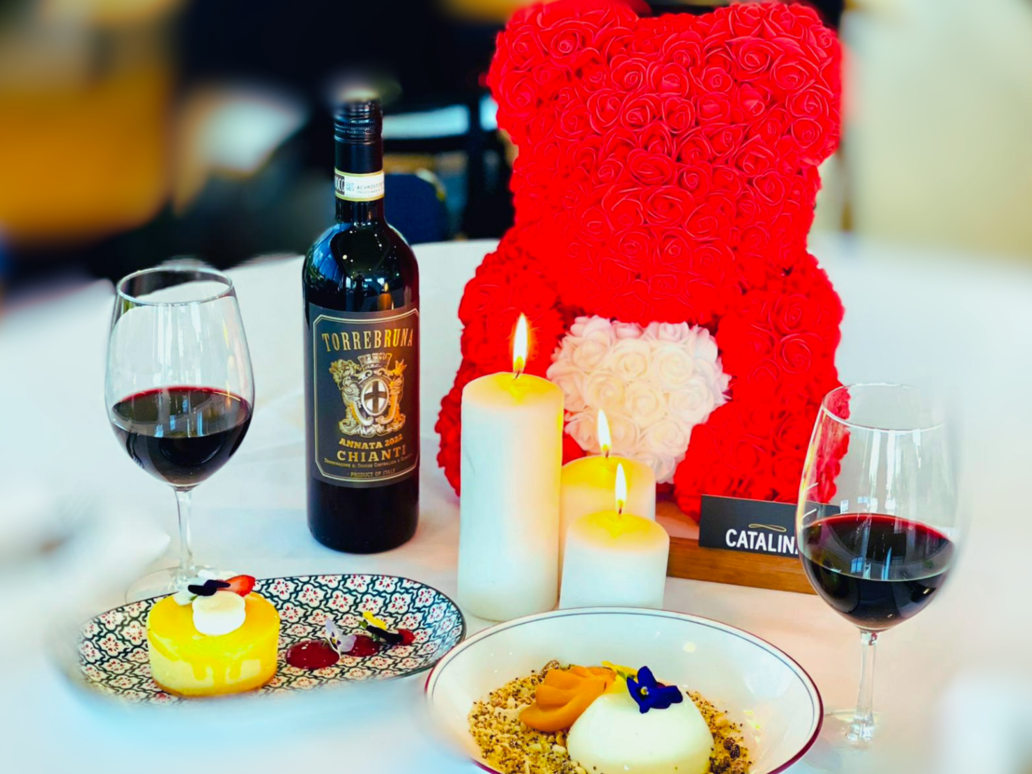 A romantic table setting with two glasses of red wine, a bottle of Chianti wine, a large heart-shaped floral decoration, three lit white candles, a dessert plate with a small cake topped with edible flowers, and a decorated plate with a panna cotta a