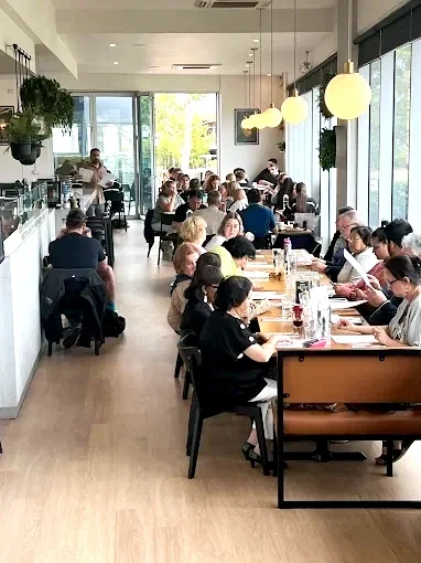 Busy cafe with customers seated at tables, large windows, modern decor, hanging pendant lights, and greenery.