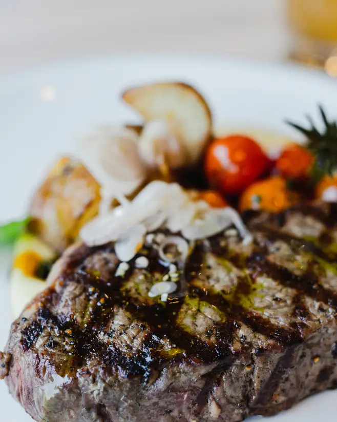 A grilled steak topped with sliced onions, served with roasted cherry tomatoes and a lemon wedge.