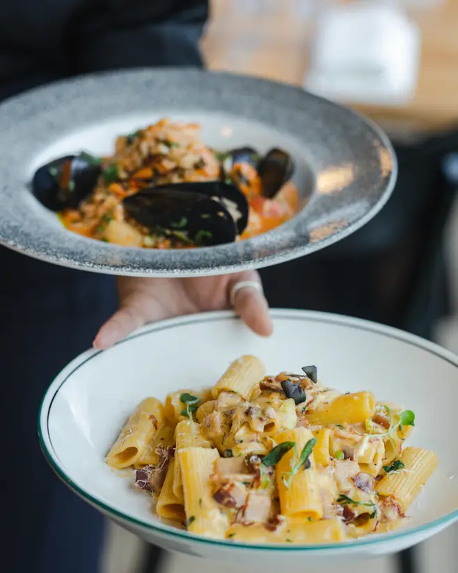 Two hands each holding a plate of pasta, with the top plate containing mussels in sauce and the bottom plate containing tube-shaped pasta with sauce and garnished with herbs.