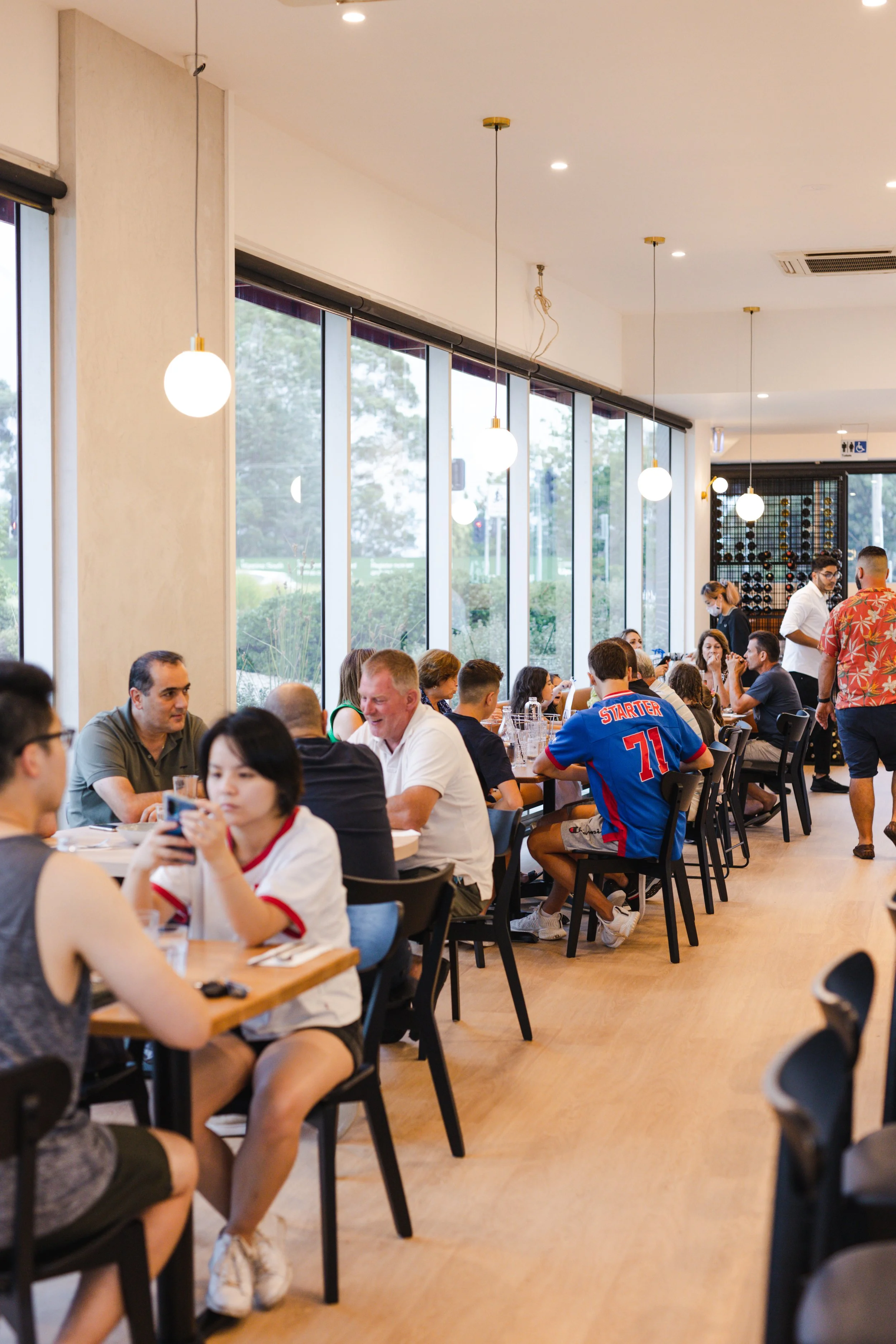 A busy restaurant with large windows and hanging pendant lights, filled with people dining and socializing. Best breakfast in knox and best dinner in knox .