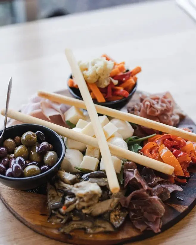 A round wooden platter with various cheeses, olives, cured meats, and sliced vegetables arranged with breadsticks