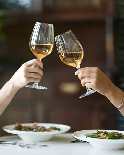 Two people clinking glasses of white wine over a table with plates of food.