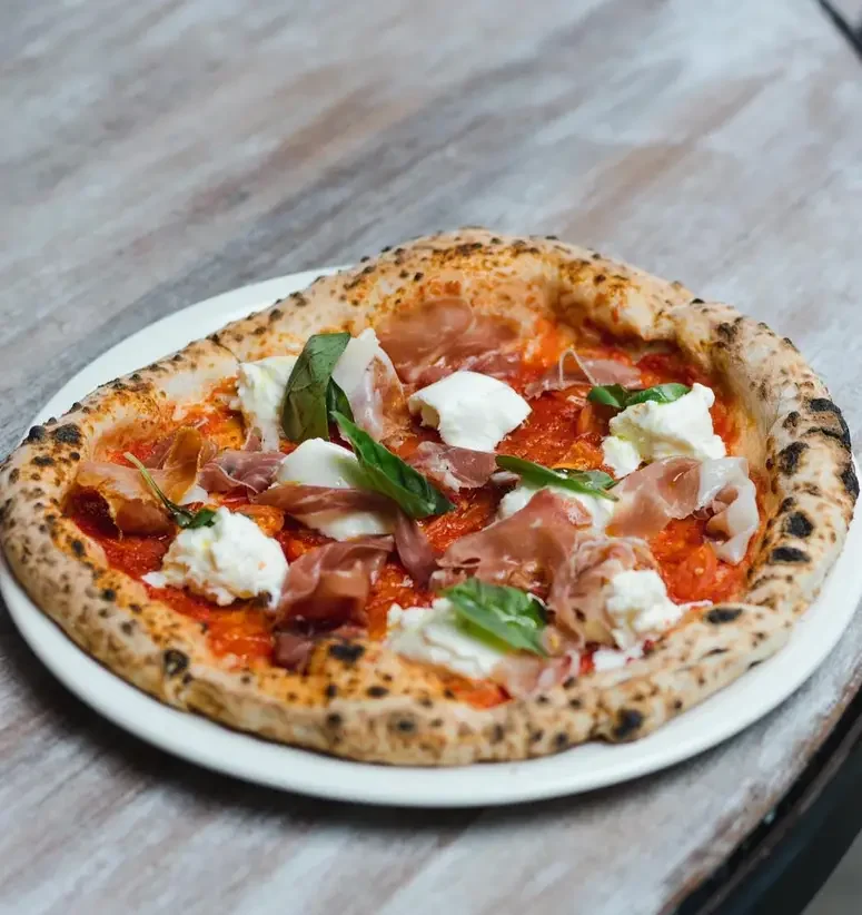 A pizza with pepperoni, prosciutto, fresh mozzarella, and basil on a wooden table.