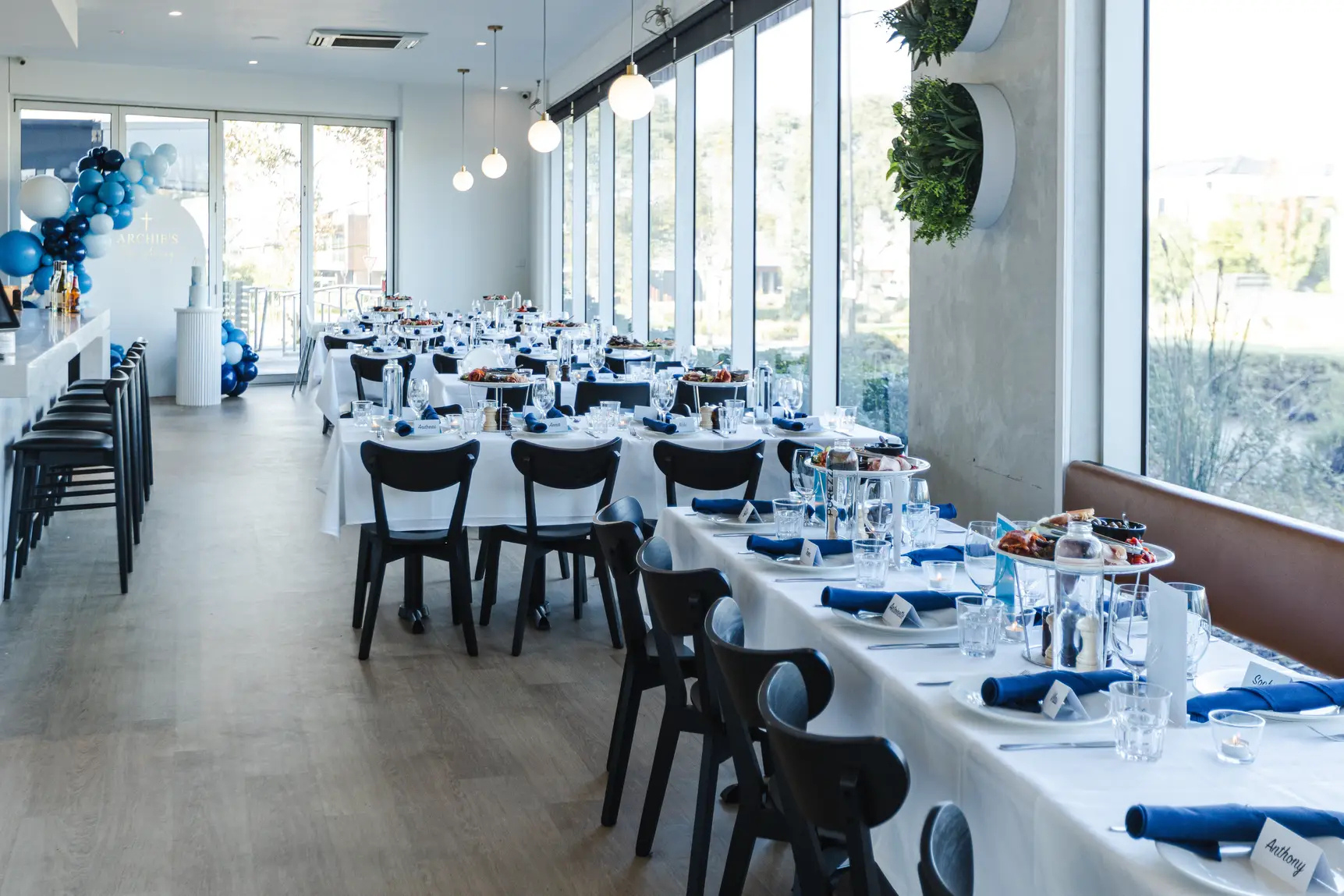 Elegant indoor dining area decorated for a celebration with white tablecloths, blue napkins, and glassware, large windows letting in natural light, and a balloon arrangement in blue, black, and white near the entrance.
Function room in knox