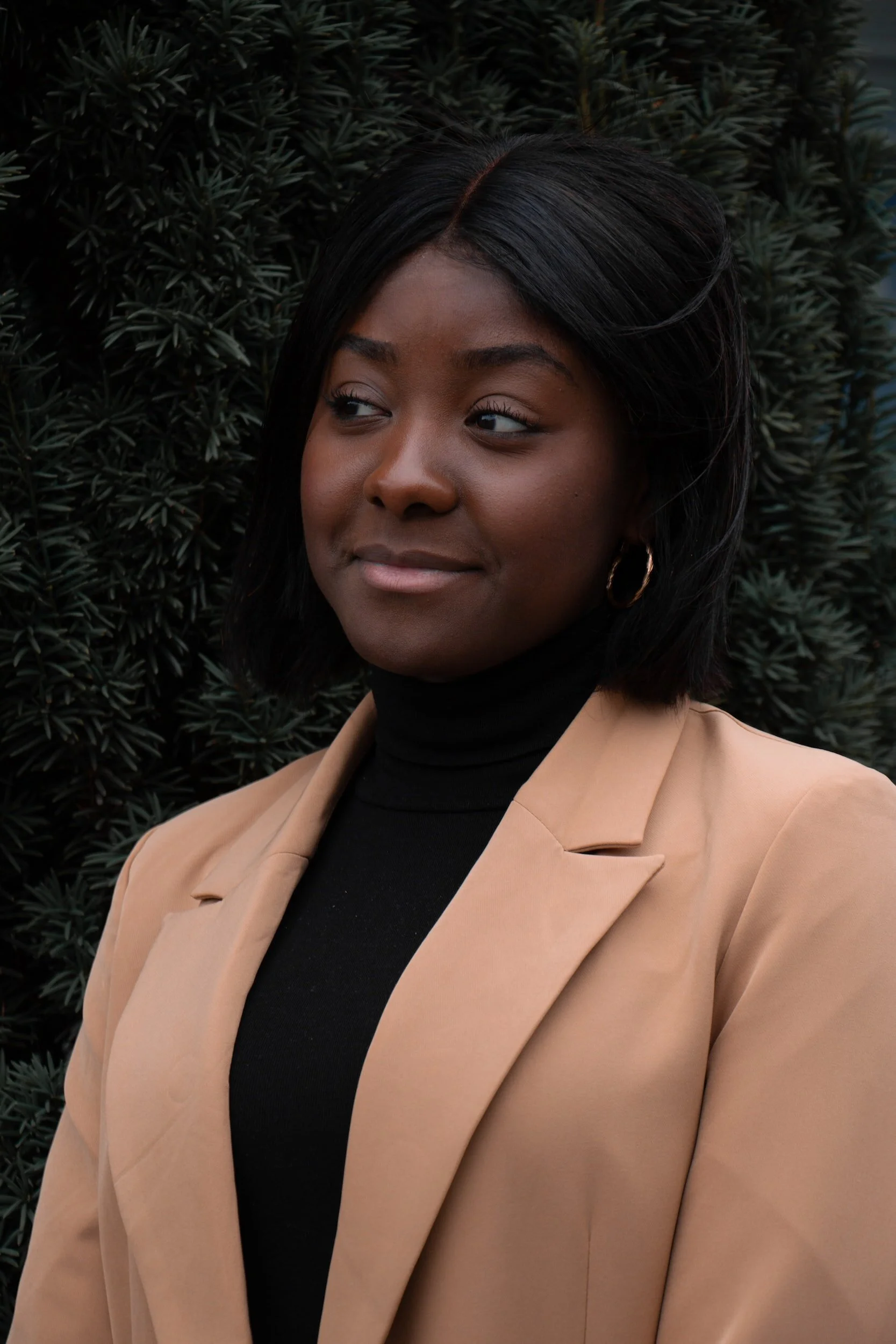 A young Black woman with shoulder-length black hair, wearing a tan blazer over a black turtleneck, standing outdoors in front of green foliage.