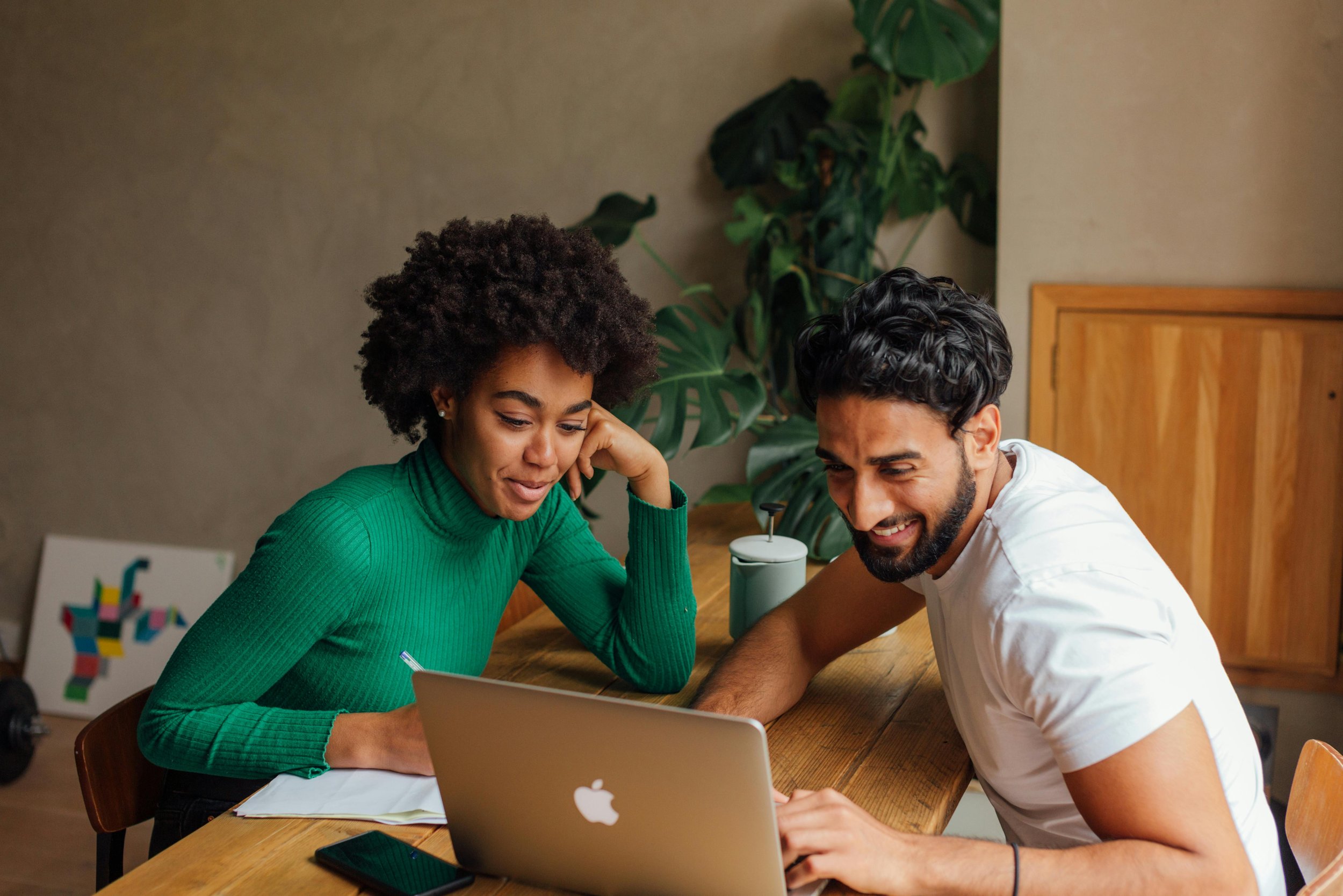 Black woman with curly afro in green sweater and South Asian man with short curly hair and beard happily looking at a laptop at a wooden table.