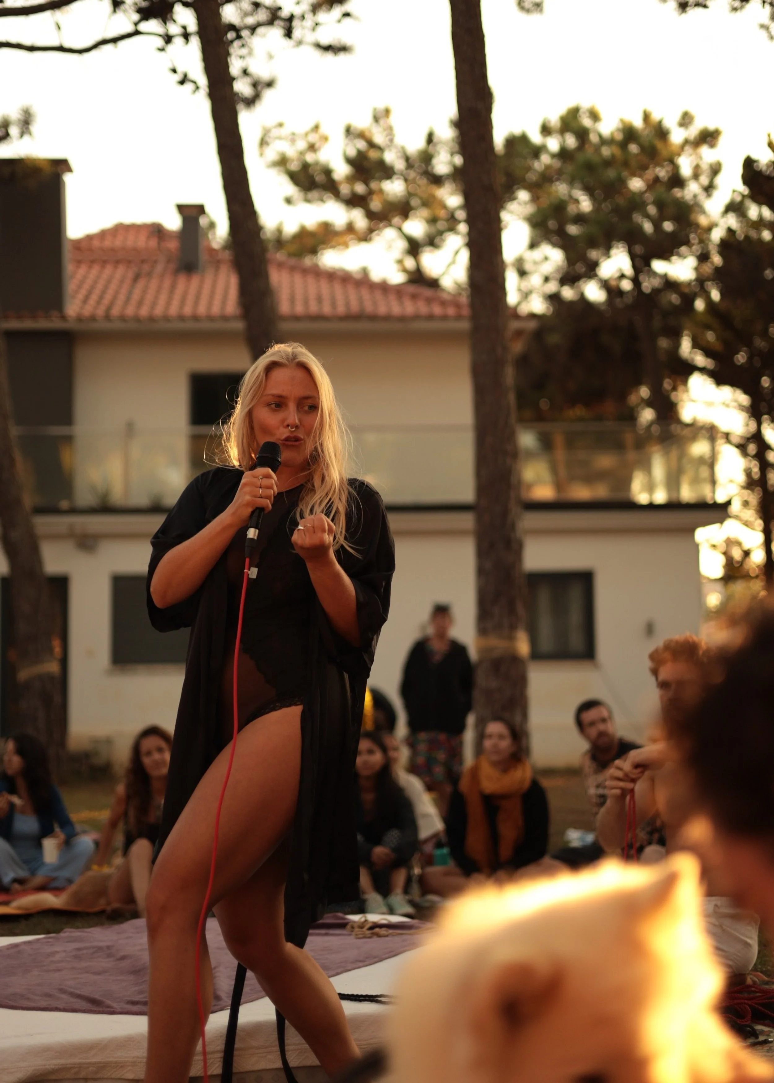 Blond facilitator with microphone in an outdoor, summery event location whilst group listening attentively. Trees in the background and colors fall golden onto the hair of the female facilitator as the sun sets sun is not visible).