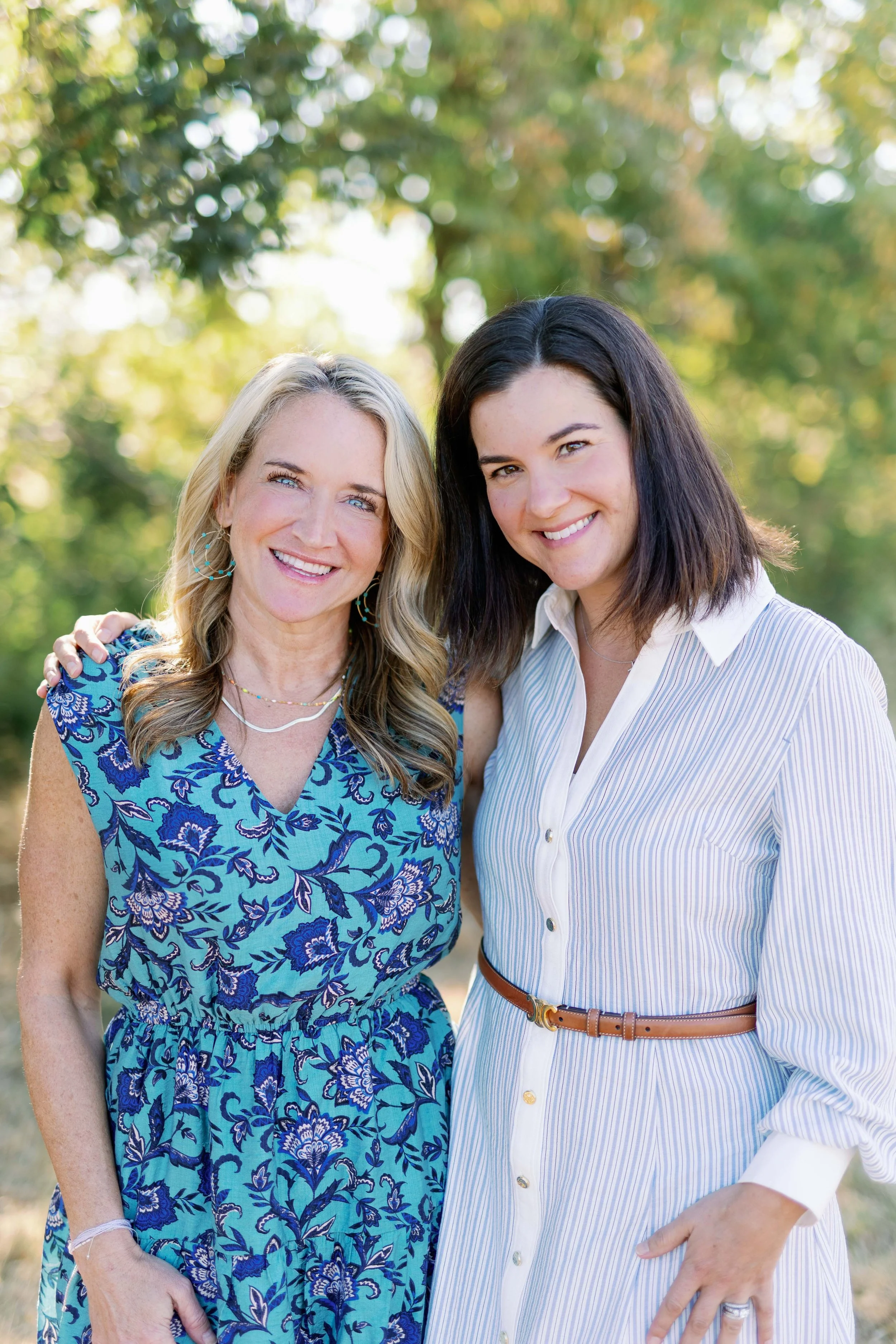 Two women smiling outdoors with trees in the background; one wearing a blue floral dress, the other in a white and blue striped dress with a brown belt.