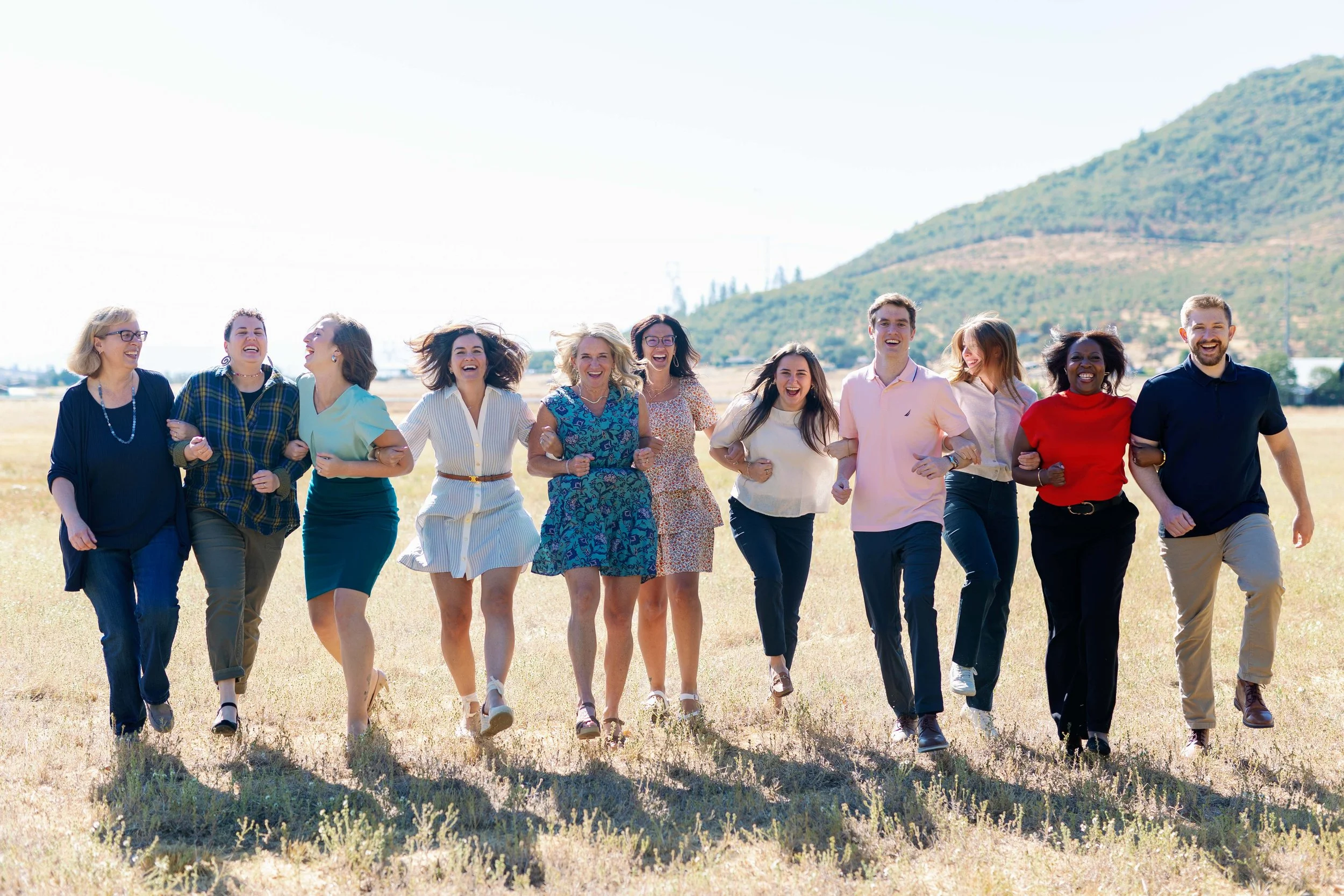 A group of diverse people holding hands and walking together outdoors on a sunny day, with a mountain in the background.