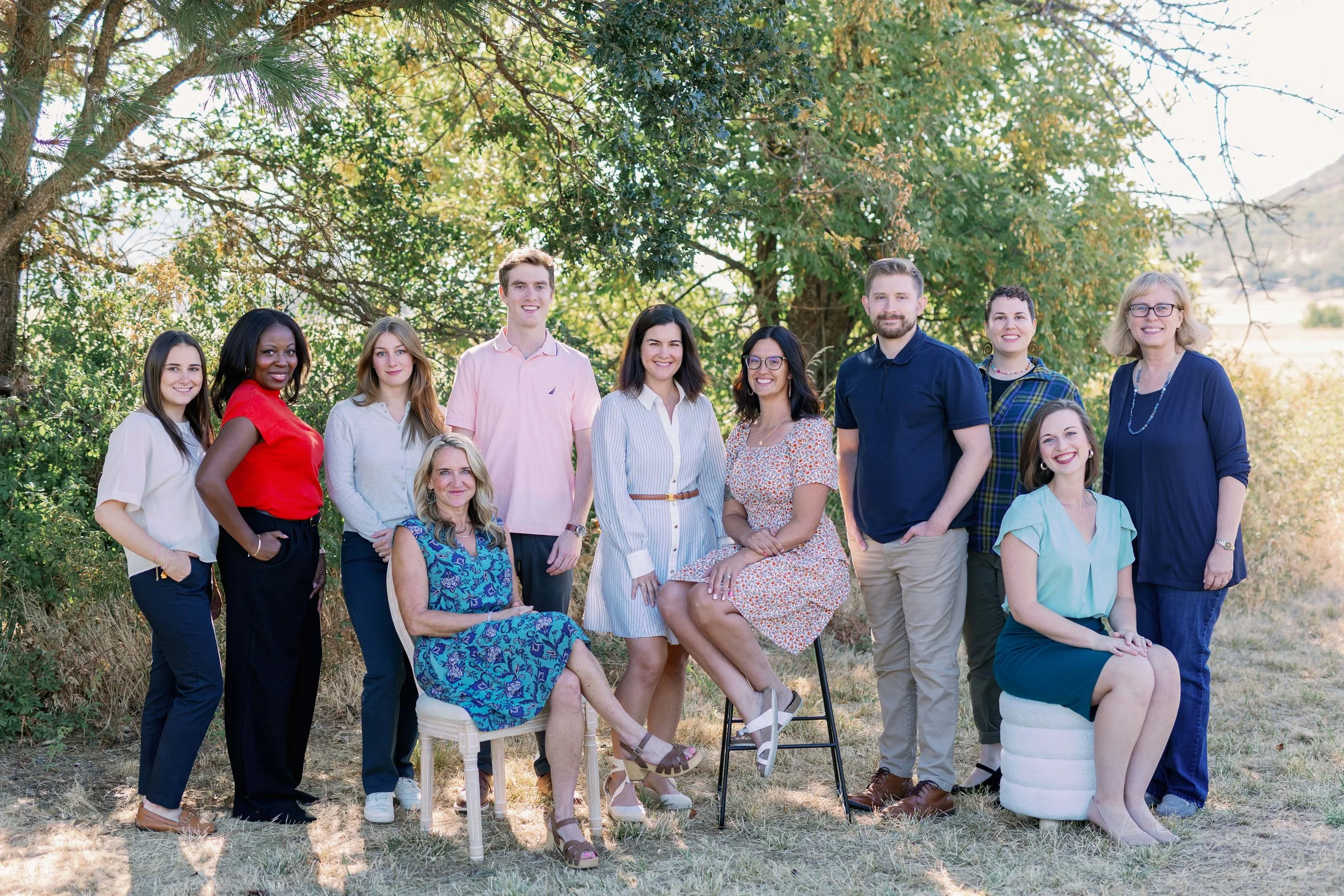 A diverse group of thirteen people, mostly women, posing outdoors under a large tree with green leaves, in a rural area with hills in the background, during daytime.