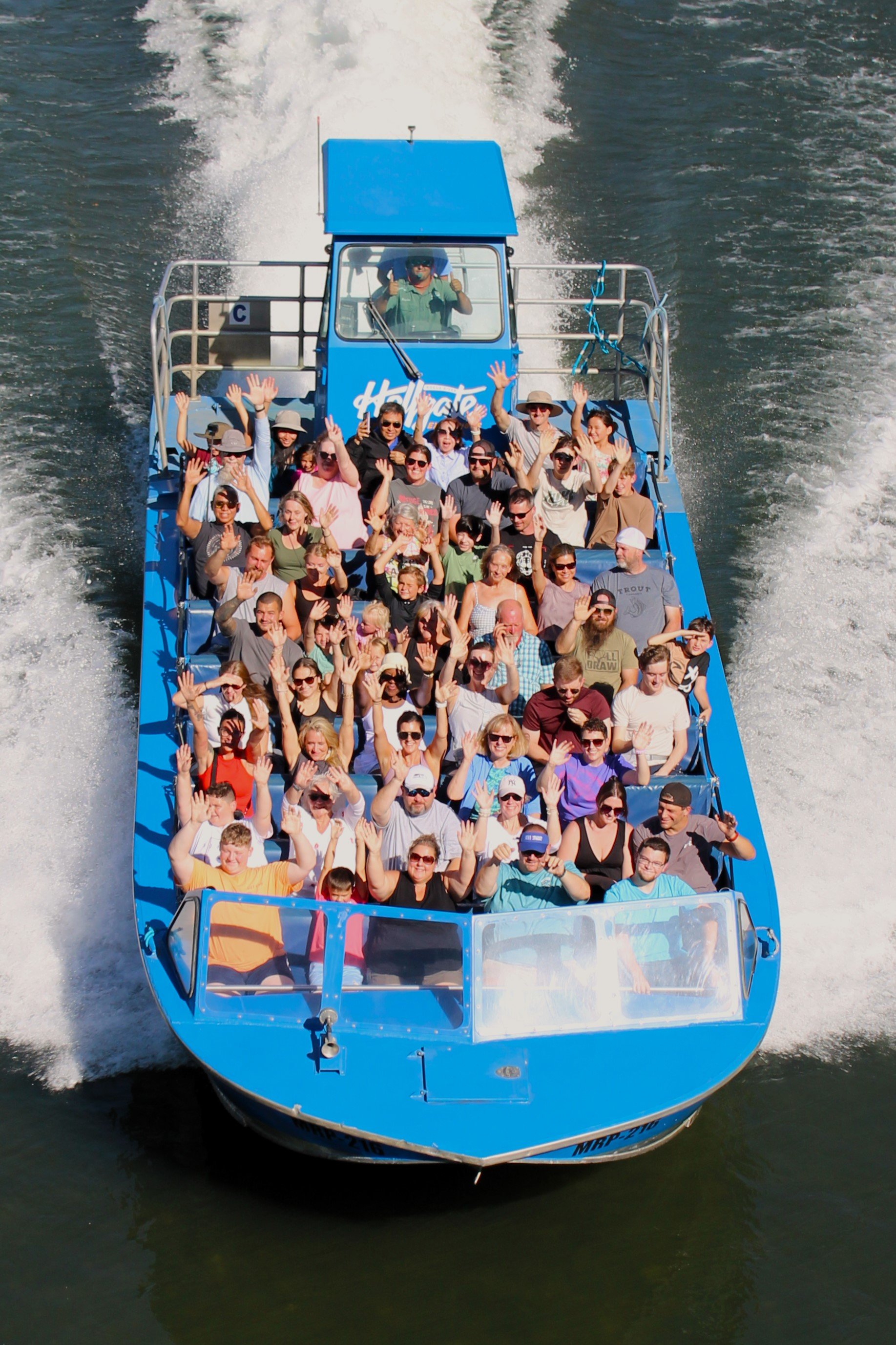 A blue boat filled with people waving and smiling, moving quickly on the water.