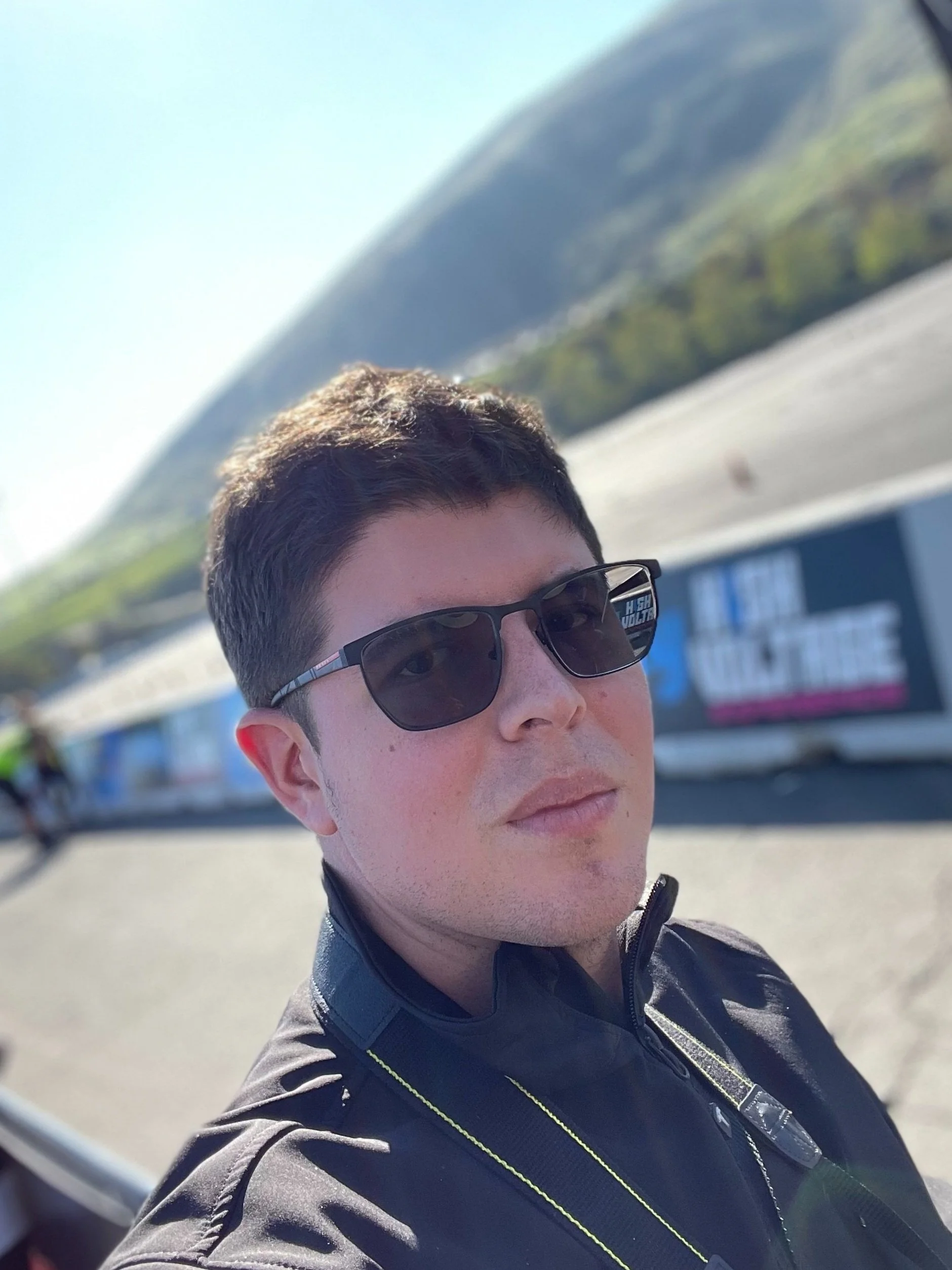A young man wearing sunglasses and a black jacket taking a selfie outdoors with a hilly landscape and a mountain in the background.