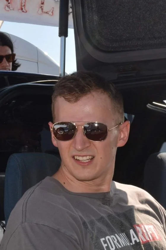 A young man with short hair and sunglasses is sitting inside a vehicle, smiling. There is another person partially visible in the background.